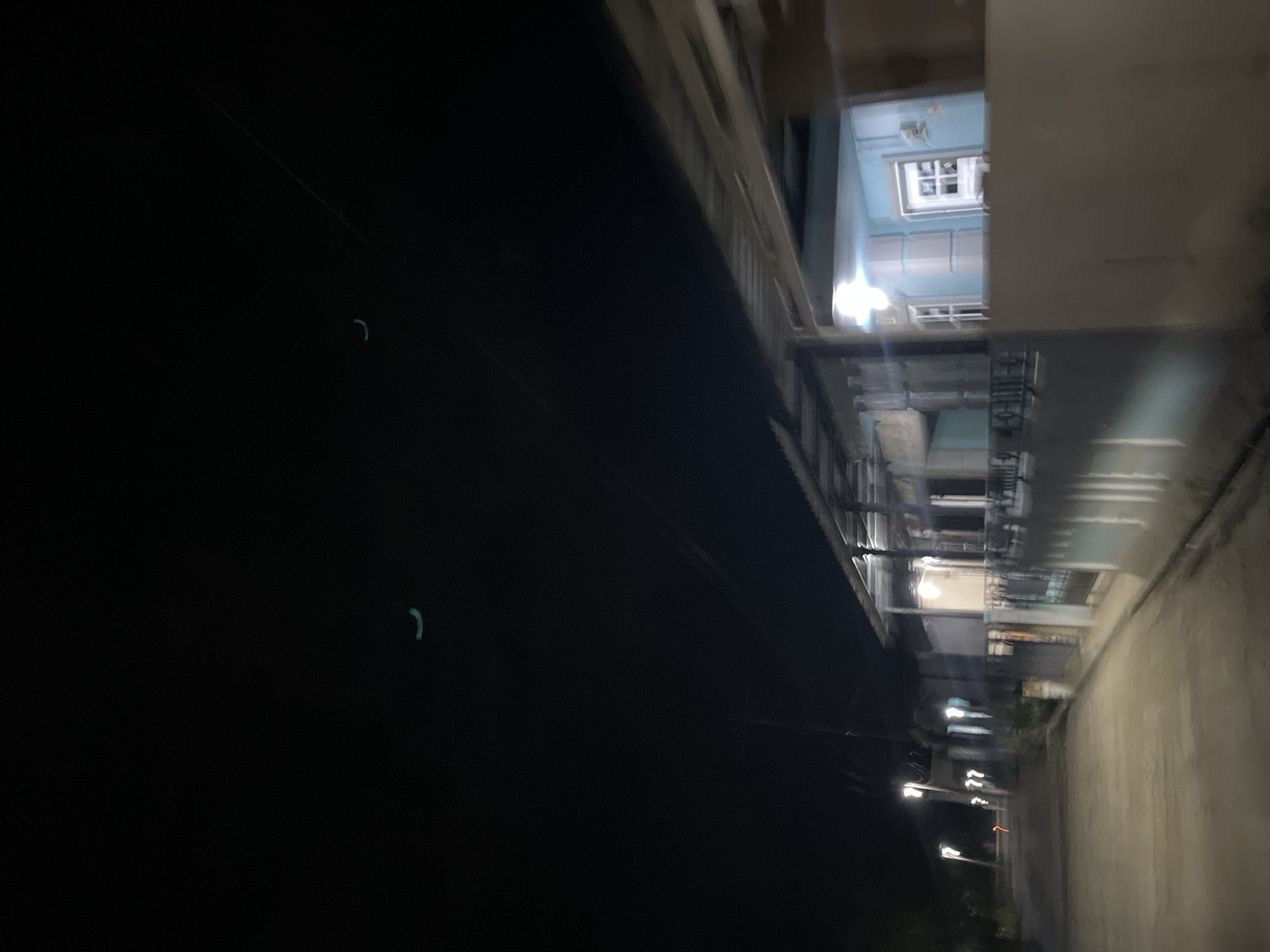 The image depicts a blurred, nighttime scene in Menanggini, Indonesia. The perspective is tilted, giving a sense of motion or an unusual viewpoint. On the right side of the frame, a paved road curves away into the distance. Along the left side of the road, a building or structure is visible. This structure appears to be multi-story, with illuminated windows and balconies, suggesting it is a residential or commercial building. The upper floors of the building are brightly lit, with one window clearly showing a checkered pattern.
To the left of the road and building, a large, dark area dominates the frame. This could represent a body of water, a park, or simply the unlit side of a street. Faint, curved lines of light are visible in this dark area, possibly reflections or distant lights. Streetlights are also present along the road, casting pools of light and creating streaks due to motion blur.
The overall mood is somewhat mysterious and urban, characterized by the darkness, artificial lights, and the blurred details. The time of day is clearly night, and the weather is not discernible from the image. No text is visible. The scene appears to capture a fleeting moment, possibly from a moving vehicle, adding to the dynamic yet indistinct quality of the photograph.