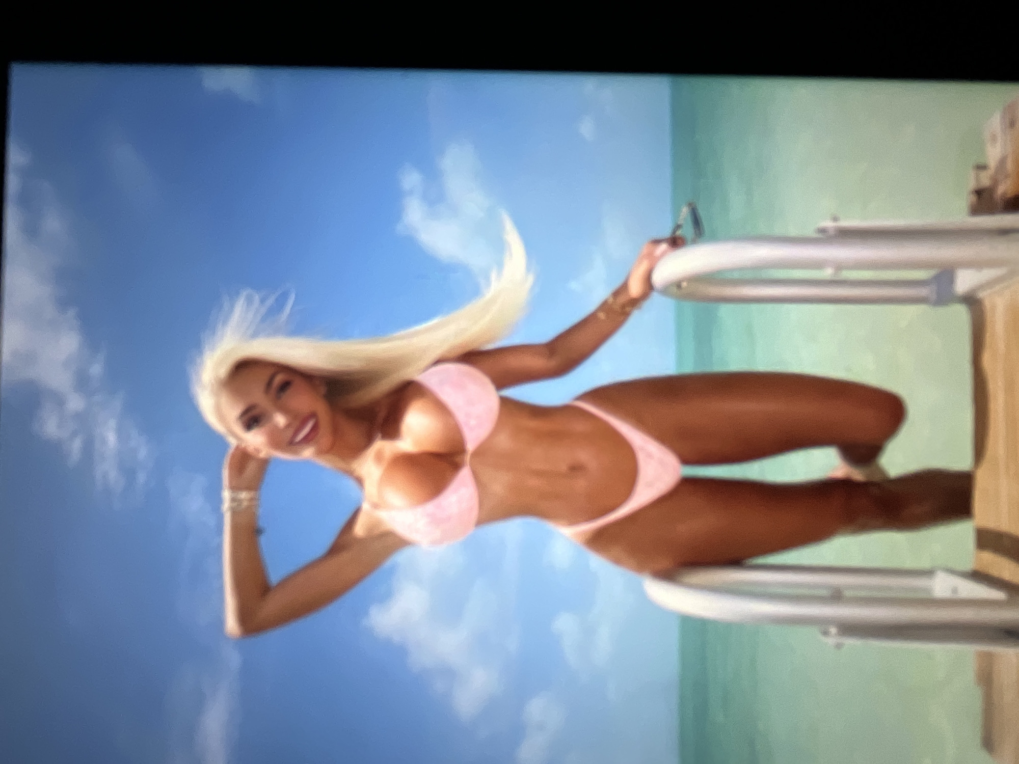 A blonde woman in a pink bikini stands on a wooden dock with a ladder leading into the turquoise ocean. She has her right arm raised and her hand behind her head, while her left hand rests on the ladder. The sky is a clear blue with a few white clouds. The image is a full-body shot of the woman, with the ocean and sky as the background. The location is not in Sofia, Bulgaria.