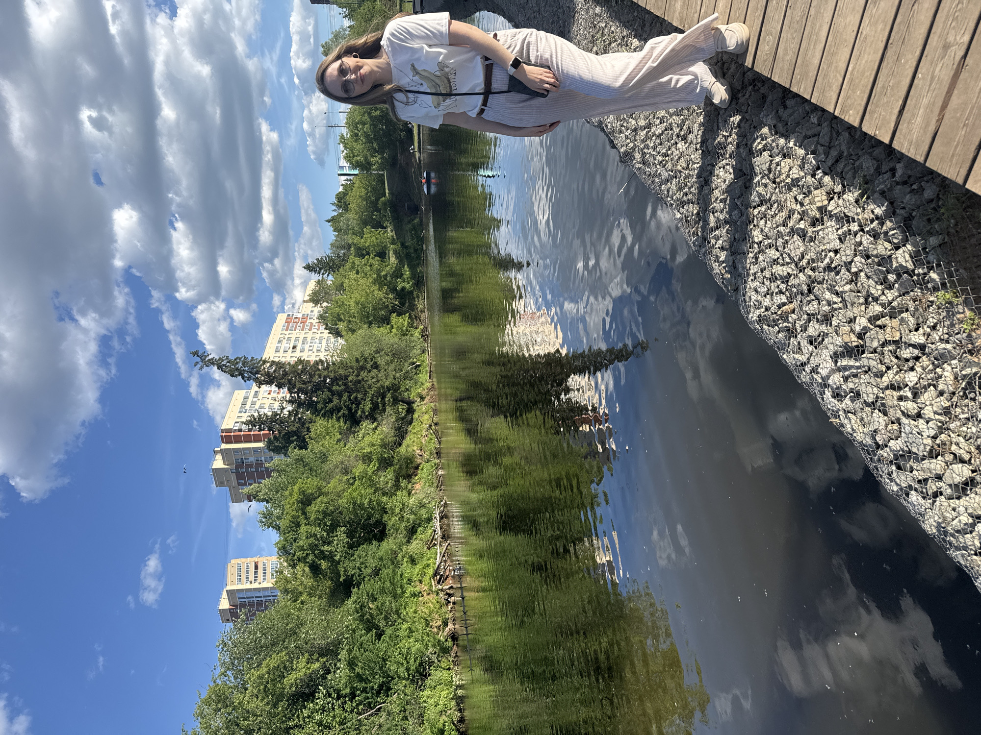 This image captures a wide-angle outdoor scene on a bright, sunny day in Izhevsk, Russia, featuring a woman standing by a body of water with urban architecture in the background.
In the right foreground, a young adult woman with light brown hair and eyeglasses stands on a wooden boardwalk, slightly angled towards the camera, with a subtle smile. She is dressed in a white short-sleeved t-shirt featuring a light-colored graphic print, light beige or white wide-leg pants with thin vertical stripes, a dark brown belt, and light-colored flat shoes. A small dark crossbody bag hangs on her left side, and a smartwatch is visible on her left wrist.
To the left of the woman and extending along the water's edge, a gabion wall constructed from wire mesh filled with light grey rocks forms the bank. The calm, dark green water of a river or large pond occupies the left and central portions of the frame, showing clear reflections of the blue sky, white clouds, and dense green trees. Small ripples are visible on the water's surface.
The opposite bank is heavily forested with lush green deciduous trees and bushes. Partially submerged logs or branches extend from the bank into the water, with several small dark birds perched on them or swimming nearby. Above the treeline, several multi-story residential apartment buildings with distinctive orange-red and light beige sections are visible, characteristic of urban architecture. Overhead lines or cables span between these distant buildings.
In the mid-ground on the water, a small white pedal boat or rowboat with two occupants is visible in the distance. The sky above is a bright blue with scattered, fluffy white cumulus clouds, and two small dark bird-like shapes are faintly visible in the upper sky. Strong sunlight is evident, casting distinct shadows from the woman and the gabion wall onto the boardwalk.