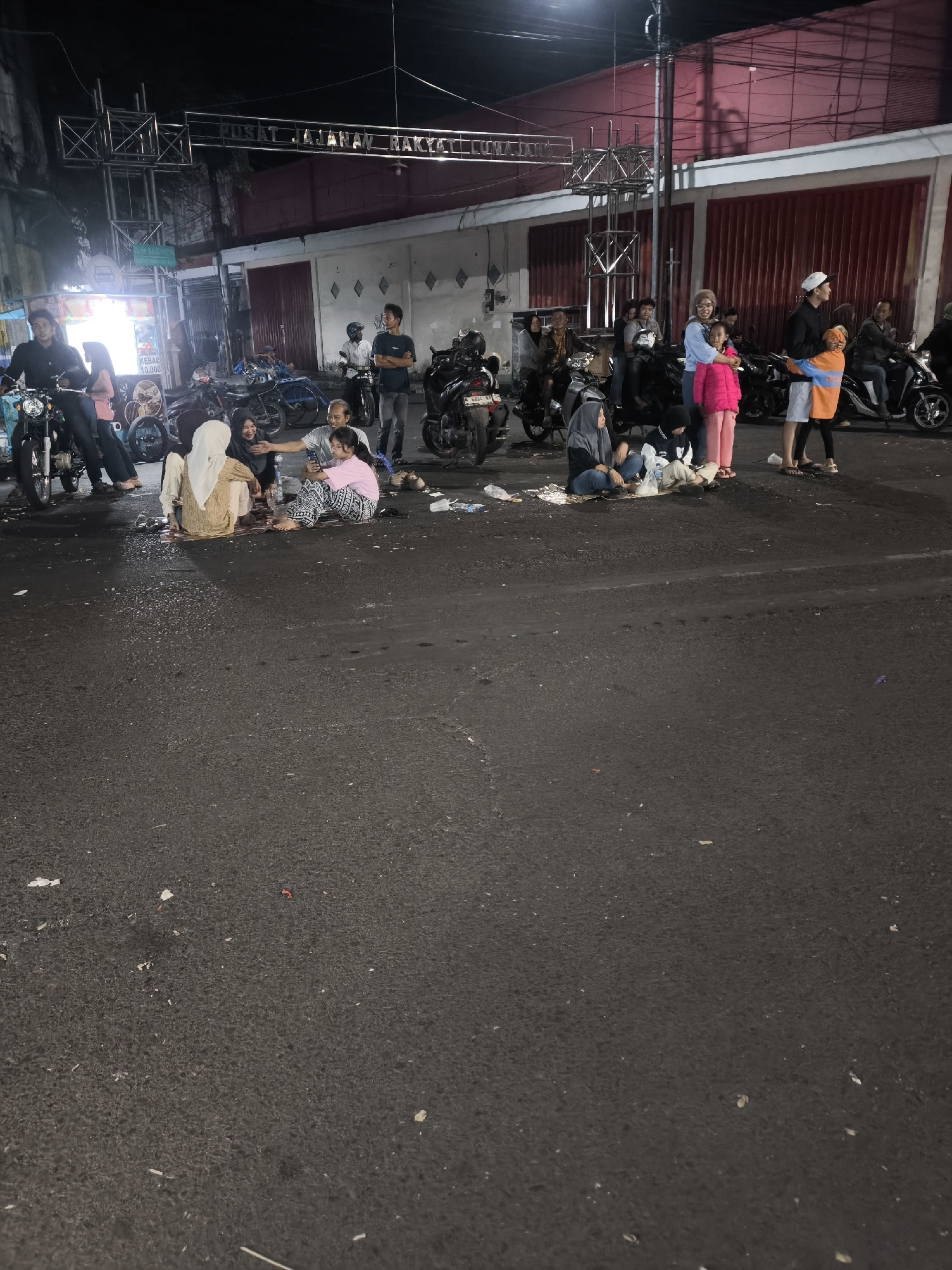 The image depicts a street scene at night in Lumajang, Indonesia. A group of people are gathered in the foreground, seated on the asphalt, some of whom appear to be eating or socializing. Several motorcycles are parked in the background, along with what appears to be a food stall. Above the scene, a sign is visible, with text that reads "PUSAT JAJANAN RAKYAT LUMAJANG". The environment appears to be an urban street with buildings and streetlights.