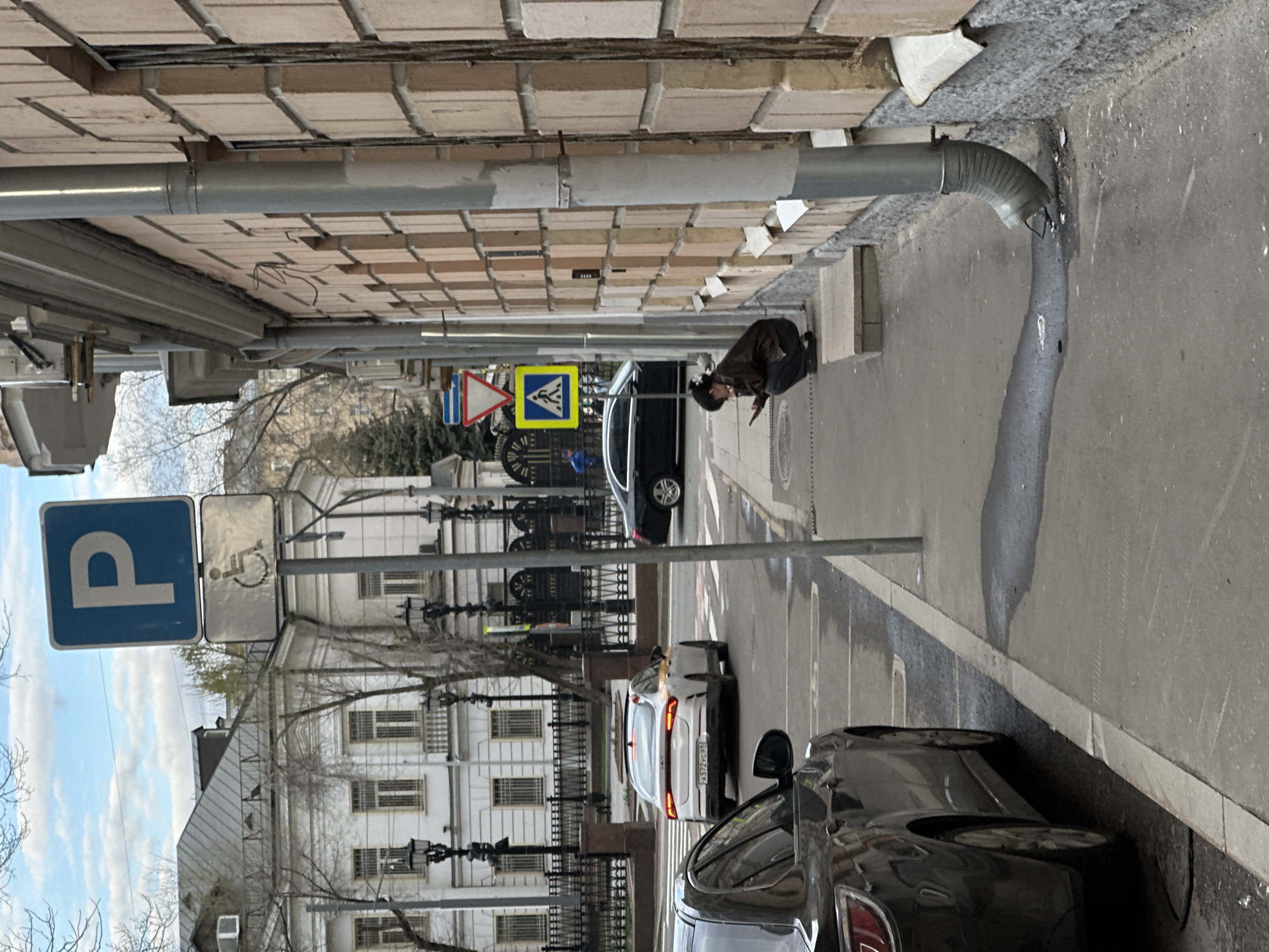 The image depicts a street scene in Moscow, Russia. A woman is crouching on the sidewalk near a black car, possibly looking at something on the ground. In the foreground, there is a parking sign with a wheelchair symbol, indicating accessible parking. The street is lined with parked cars, including a white car with a license plate visible. In the background, there is a building with multiple windows and architectural details. Overhead, a drainpipe runs along the edge of a building. There are pedestrian crossing signs indicating a pedestrian area, and the overall environment suggests a typical urban street.