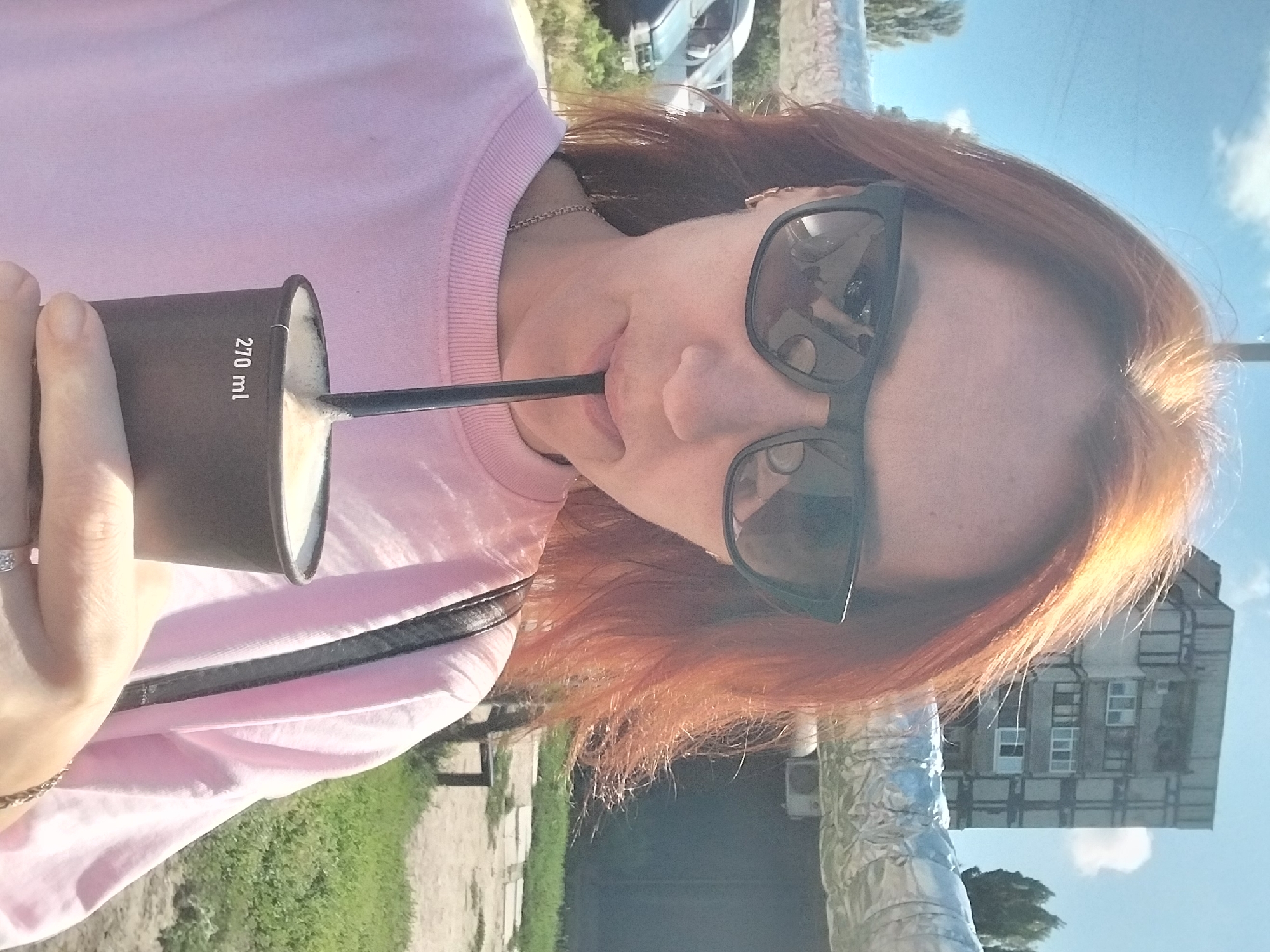 A woman with auburn hair and sunglasses drinks from a black paper cup with a straw. She wears a pink sweatshirt and a silver necklace. In the background, there is a grey apartment building, a silver pipe, trees, and a blue sky with clouds. The scene appears to be in an urban outdoor setting, possibly in Dnipro, Ukraine, given the architecture and general environment.