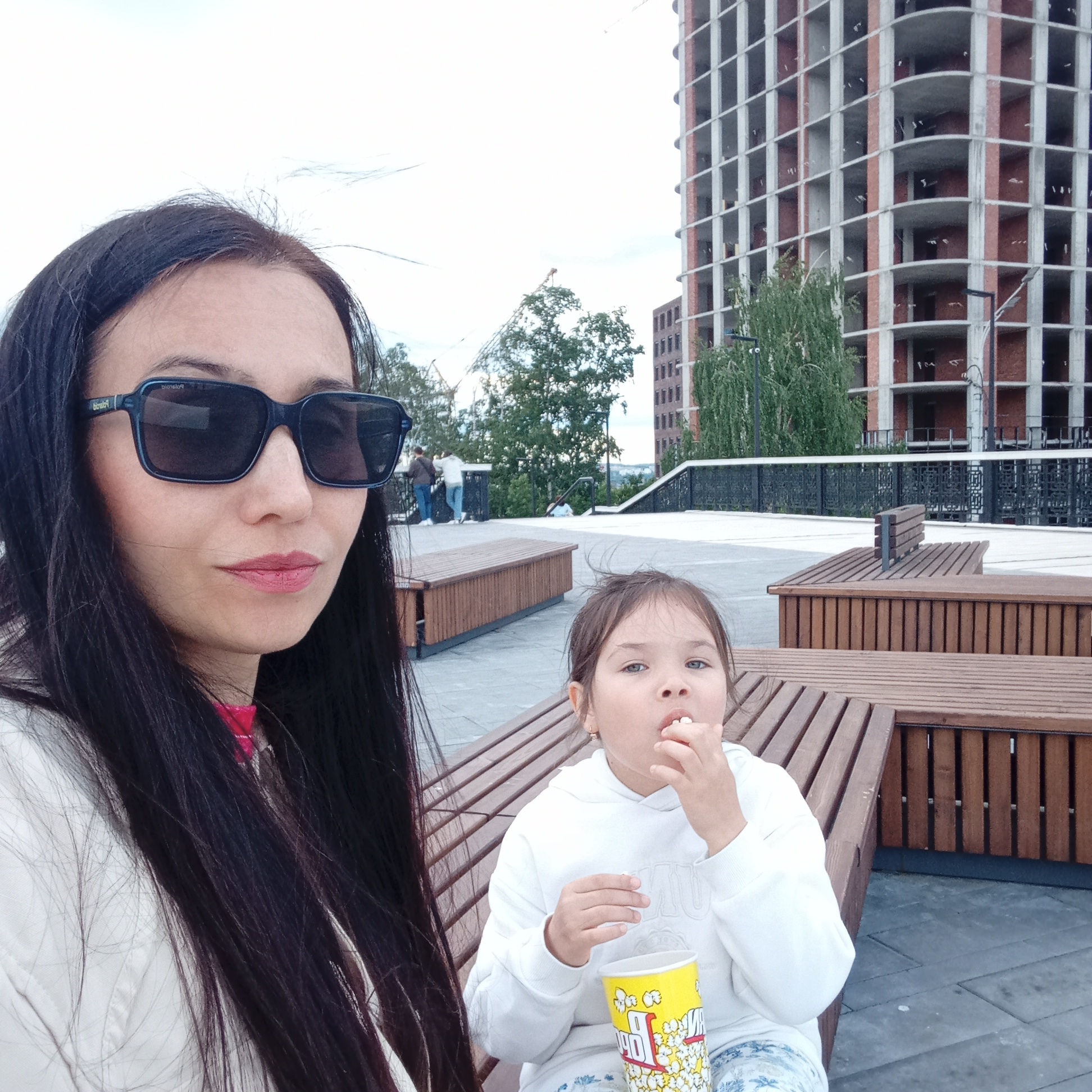 A medium-shot image depicts a woman and a young girl in an outdoor urban public space in Izhevsk, Russia, under an overcast sky.
In the left foreground, a woman with long, dark, straight hair and fair skin is shown from the chest up, wearing dark rectangular sunglasses with "Polaroid" branding visible on the left temple. She wears a light-colored outer garment, possibly white, over a bright pink inner layer visible at her collar. Her lips are painted with a pinkish-red lipstick, and she gazes directly at the camera.
To her right, a young girl, approximately 3-6 years old, is seated on a dark brown wooden slatted bench. She has light brown, slightly disheveled hair and blue eyes. She is actively eating popcorn, holding a piece to her open mouth with her right hand, while her left hand supports a yellow popcorn cup with red "POPCORN" branding. The girl wears a white hooded sweatshirt with subtle light-colored detailing on the front and light-patterned trousers.
The foreground and mid-ground are characterized by modern, dark brown slatted wooden benches arranged on light-grey paved ground. A dark metal railing with decorative elements spans horizontally in the mid-ground. The background features a tall, multi-story building under construction or unfinished, identifiable by its exposed concrete structural grid with brick infill and a distinctive curvilinear corner. Green trees are visible, particularly between the railing and the large building, and further left. Several indistinct figures are present on a raised platform in the far background. The overall lighting is diffused, consistent with a cloudy day.