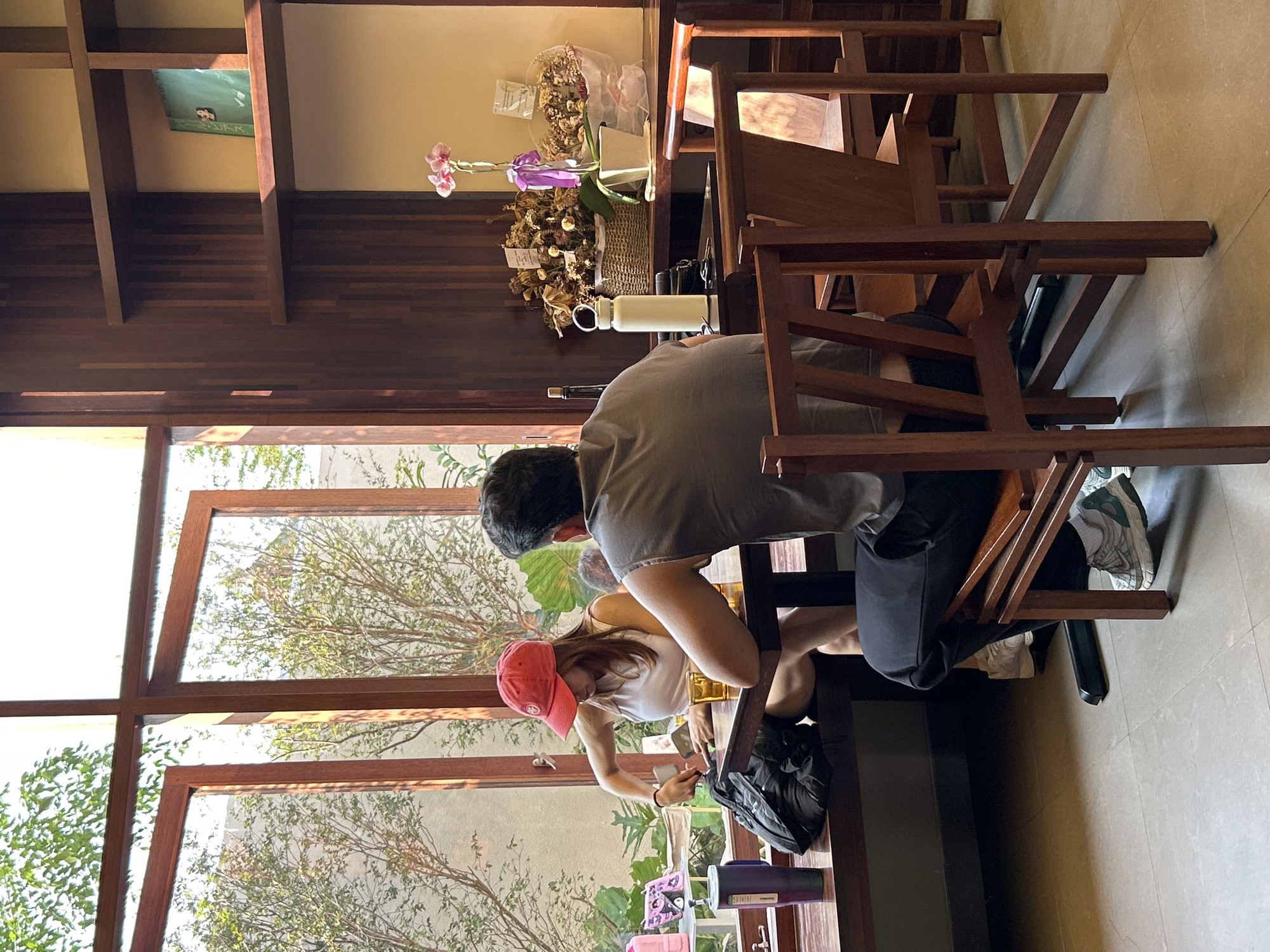 A woman wearing a red baseball cap and white tank top is sitting at a dark wooden table in what appears to be a cafe in North Kuta, Indonesia. She is facing away from the camera and is rummaging through a black backpack. A man in a grey t-shirt is seated opposite her, his back to the camera. A large window with wooden frames looks out onto lush greenery. On a wooden shelf to the right, a potted orchid and a basket of dried flowers are visible. The floor is tiled with a light grey material.