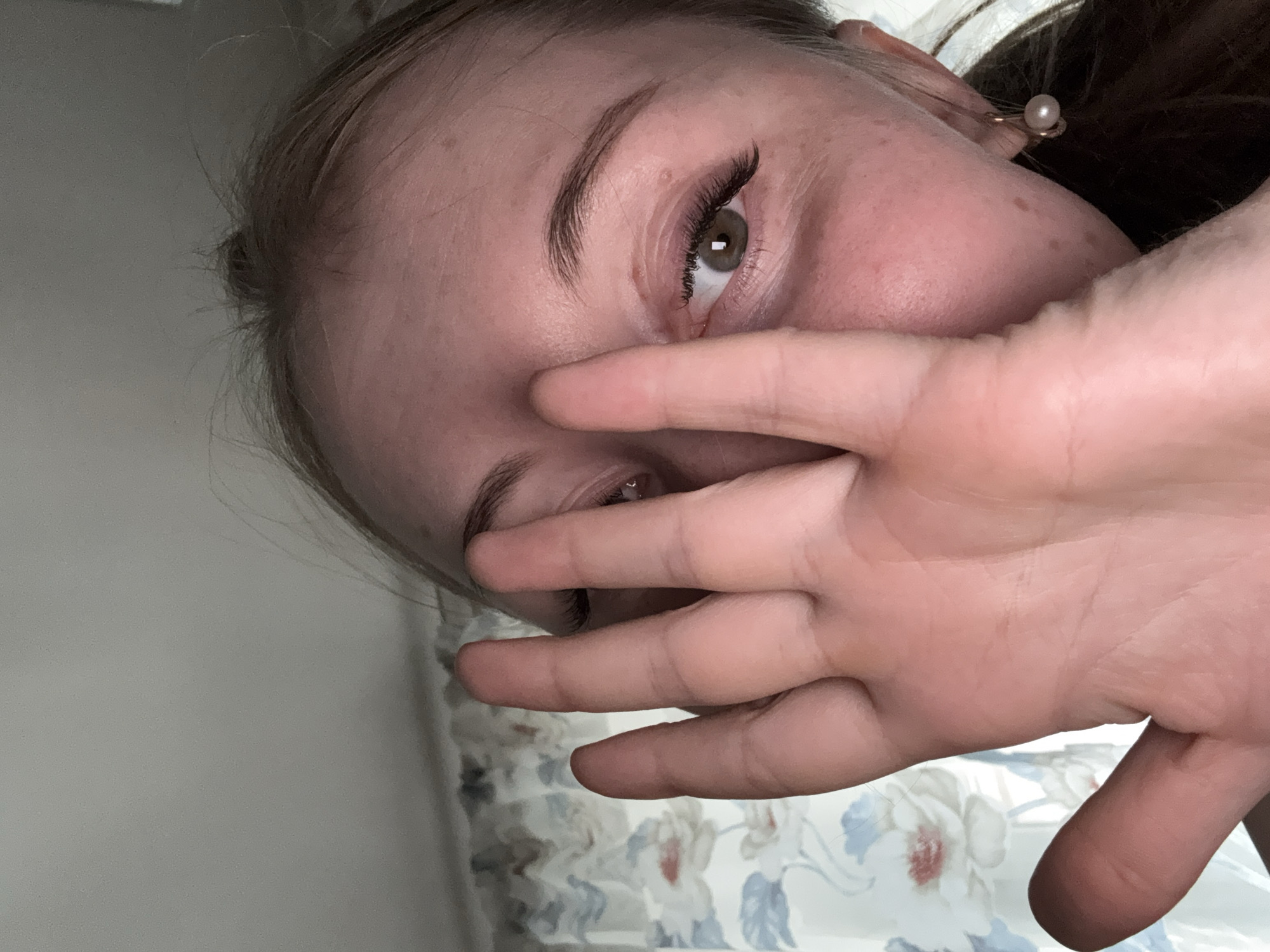 The image captures a close-up, angled portrait of a person, likely a young woman, partially covering her face with her right hand. Her head is tilted to the left, and her visible right eye, which is light brown or hazel, looks directly towards the viewer. This eye is framed by dark, long eyelashes, possibly extensions, and a well-defined eyebrow. Her fingers are spread slightly, and her palm obscures the lower portion of her face, including her nose and mouth.
Her light brown hair is pulled back, revealing her ear where a small pearl earring is visible. The skin on her forehead and cheek shows a few freckles or marks. The background on the left side is a plain, light grey surface. On the right and below her hand, a floral pattern fabric with white, blue, and red flowers on a lighter background is visible, suggesting curtains or bedding in an indoor setting. The lighting is soft and diffused, indicating an interior space, possibly during the day or under indirect artificial light. There is no visible text in the image. The pose with the hand partially covering the face could suggest shyness or a playful gesture.