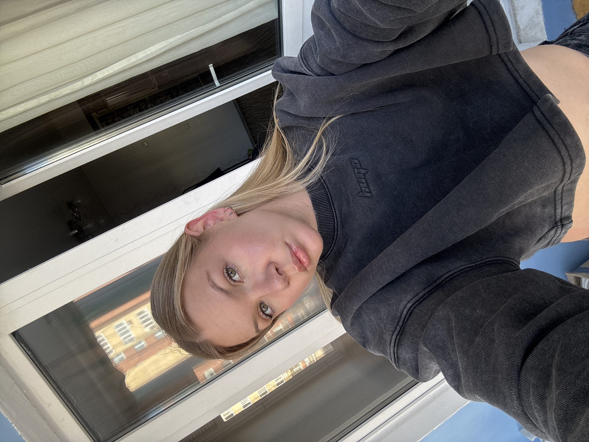 A young woman is taking a selfie indoors. She has blonde hair, fair skin, and is wearing a dark grey sweatshirt with a subtle logo. The background features a window through which a building with a distinct yellow and orange facade, common in the city of Perm, Russia, can be observed. The sky is visible through the window in the bottom corner.