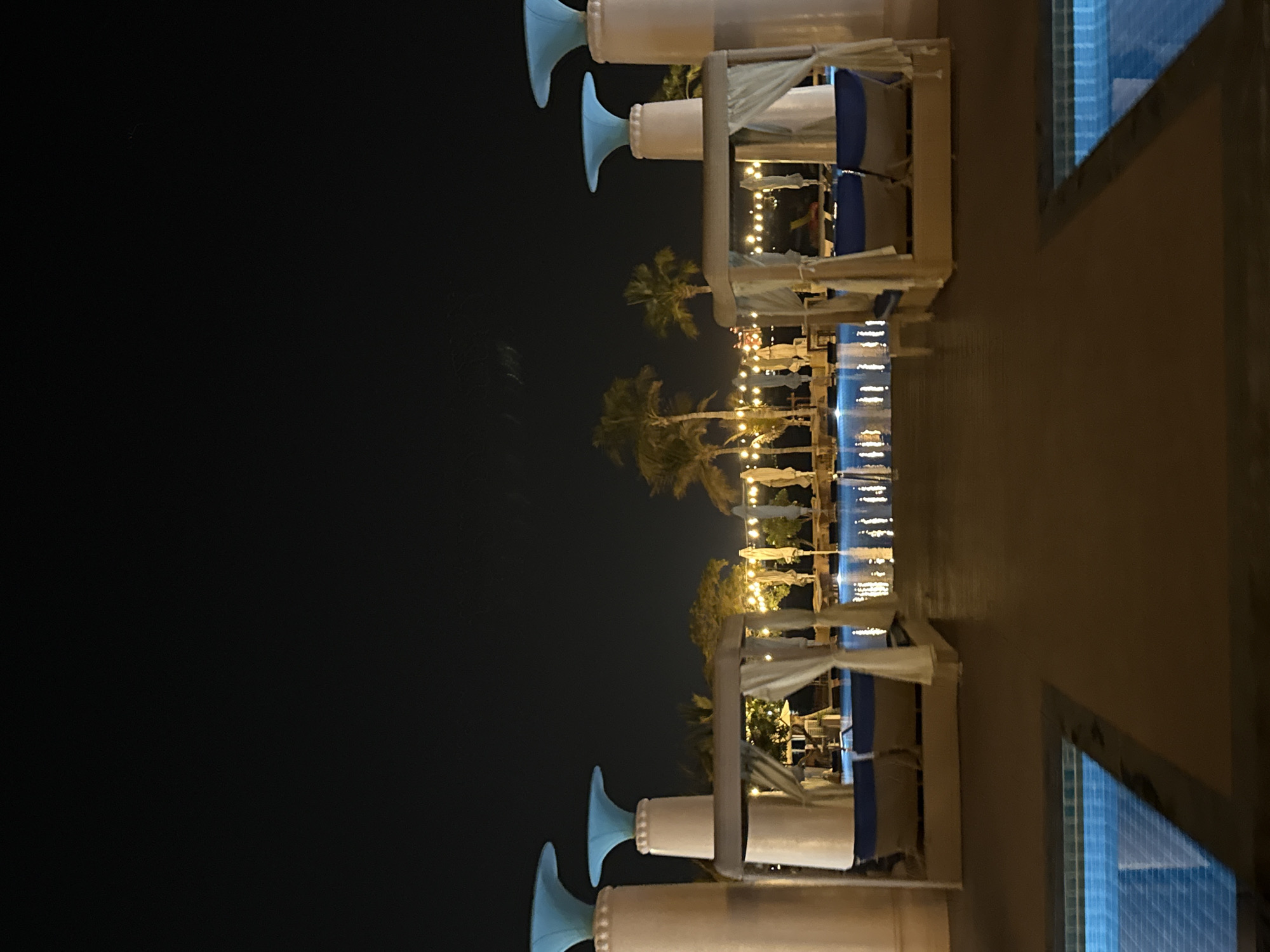 An outdoor swimming pool area is illuminated at night. Two cabanas with white curtains and lounge chairs are situated on a deck. Beyond the cabanas, a long swimming pool reflects string lights that are suspended between palm trees and poles. There are no people visible in the image. The image appears to be taken at a resort or hotel in Dubai, United Arab Emirates.