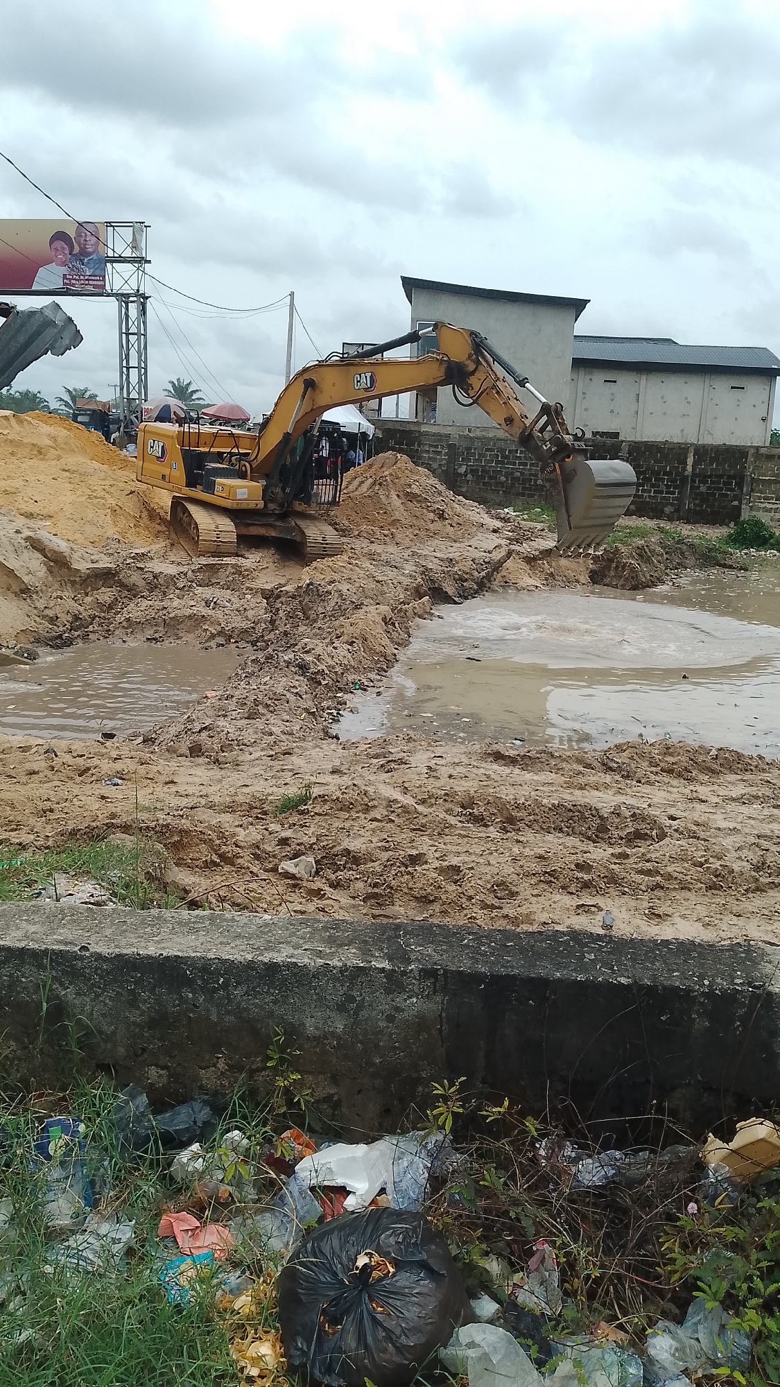 An overcast day in an urban environment. A yellow Caterpillar excavator is partially submerged in muddy water, appearing to be dredging or moving dirt. A large pile of earth is next to the excavator. In the foreground is a concrete barrier with accumulated trash and vegetation in front. A building with a flat roof and barred windows is visible in the background, along with a billboard. People can be seen near the excavator, possibly observing the operation. The location is indicated as Unknown, Unknown.