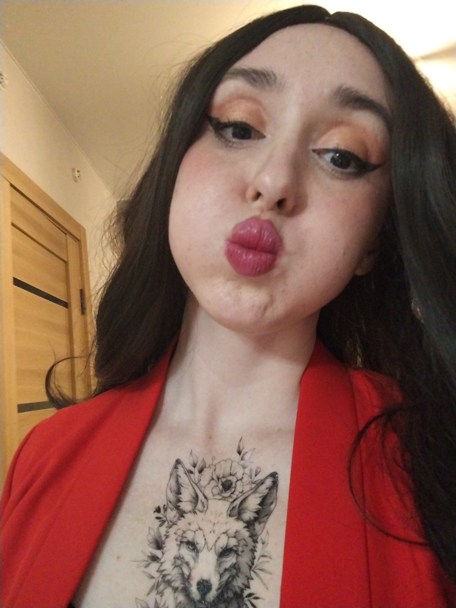 A close-up selfie of a woman. She has long, dark hair and fair skin. She is wearing a red jacket. She has a fox tattoo with floral elements on her chest. She is puckering her lips in a kiss. The background shows a light-colored wall and a wooden door. The image does not provide information to confirm the location of Izhevsk, Russia.