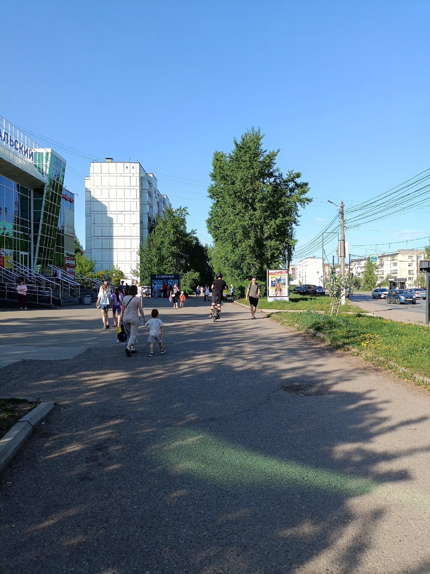 The image depicts an outdoor scene in the city of Chaikovsky, Russia, recognizable by the Cyrillic text “Чайковский” on a building facade in the background. It's a sunny day with clear blue sky.
A group of pedestrians is walking on a wide sidewalk. Among them, a woman is holding hands with a young child, and other adults and children are present in the background near the entrance of a building. In the distance, two men are seen walking along the same path, one of whom is riding an electric scooter.
Parallel to the sidewalk, a street with moving cars can be observed. A billboard stands along the roadside. Power lines stretch across the sky. Buildings of varied heights and designs make up the urban backdrop. The foreground is characterized by shadows and pavement details.