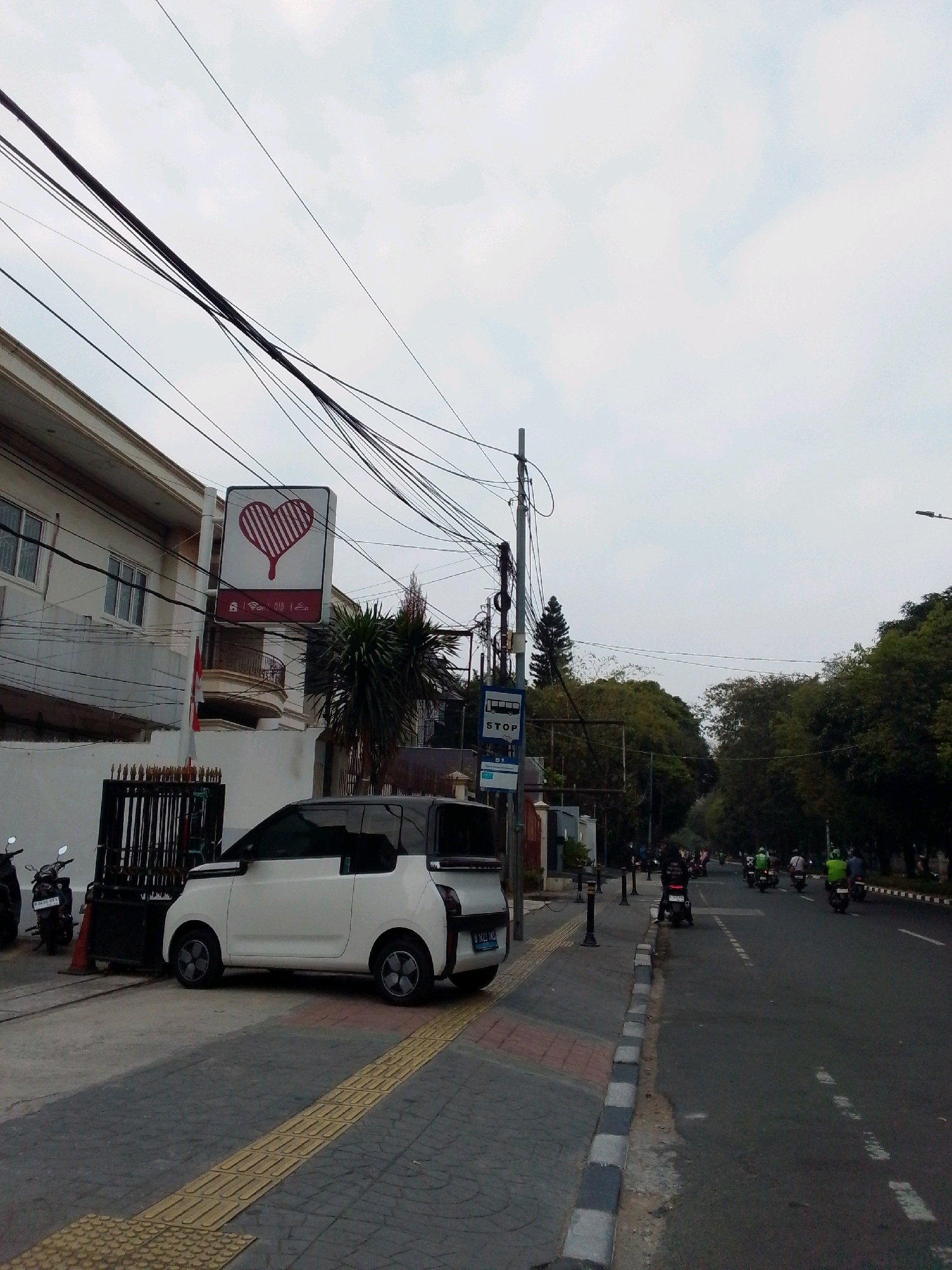 A street scene in East Jakarta, Indonesia, features a white electric car parked on the side of the road. The car is a two-door model with a black roof and distinctive gray rims. Adjacent to the car is a pedestrian walkway with tactile paving. Further down the street, a traffic light pole with a "STOP" sign is visible, indicating a potential intersection.
On the left side of the street, a building with white walls and multiple windows is partially visible, with a sign displaying a red heart shape and vertical stripes. Several power lines crisscross overhead, a common sight in urban Indonesian environments. A black metal fence and a flagpole with an Indonesian flag are also present in front of the building.
The road itself is a two-lane street with a moderate amount of traffic, primarily consisting of motorcycles. Several individuals are seen riding motorcycles, some wearing helmets and brightly colored clothing. The street is lined with trees, creating a somewhat green urban environment. The sky is overcast with scattered clouds, suggesting a muted daylight setting. The overall scene depicts a typical street view in a bustling Indonesian city.