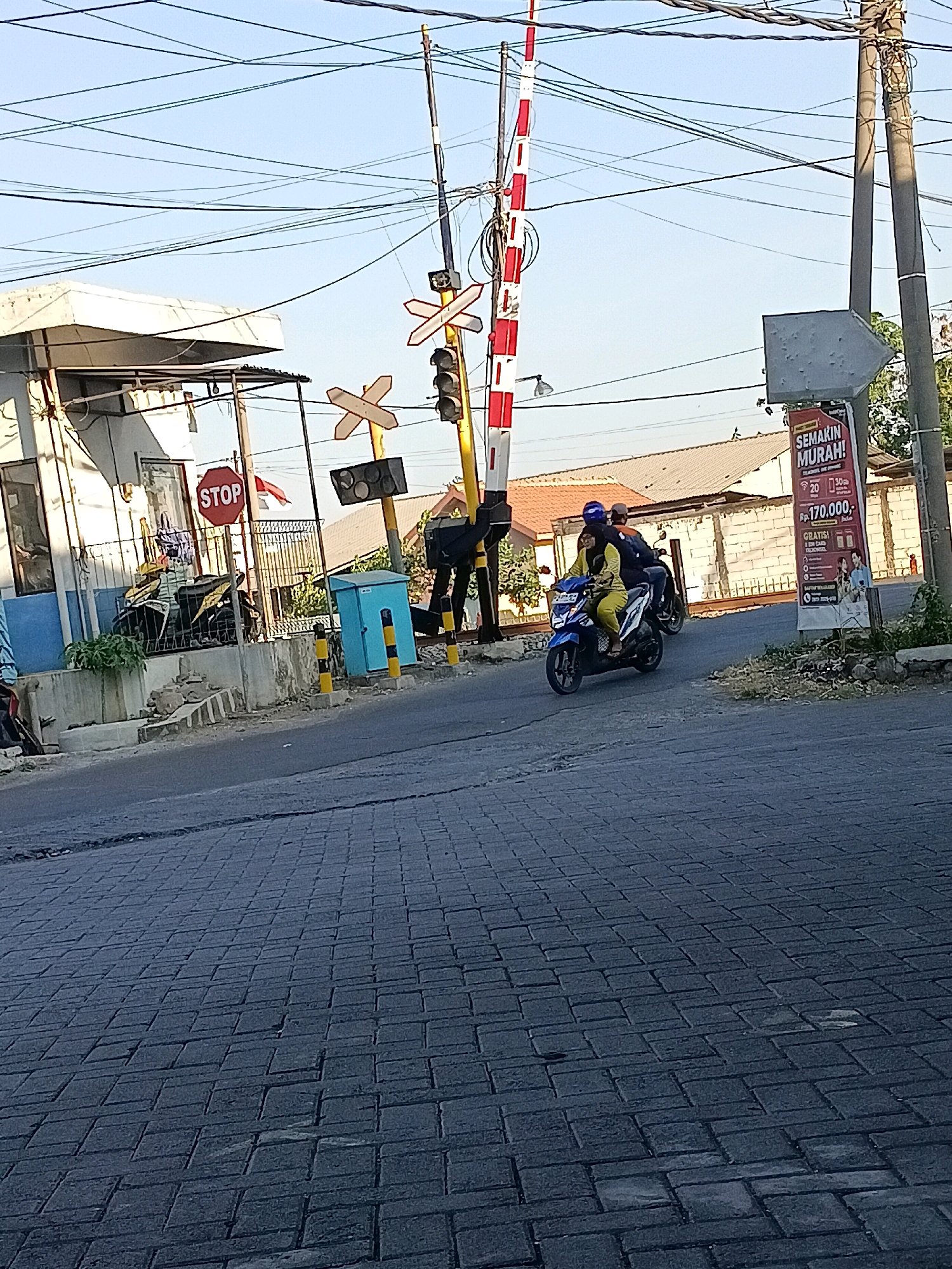 This image captures a scene at a railroad crossing in Kletek, Indonesia. In the foreground, a paved road with a patterned brick surface leads towards the crossing. Two individuals are riding a blue motorcycle, with the driver wearing a blue helmet and a passenger in yellow attire. They are approaching the crossing from the right side of the frame.
To the left of the road, there is a white building with blue trim, possibly a small office or guardhouse. Adjacent to it is a metal fence and a "STOP" sign. Further to the left, several motorcycles are parked. A small blue utility box with yellow and black striped posts is located near the curb.
The railroad crossing itself is equipped with a red and white striped barrier gate that is partially lowered, and traffic signals are visible. A tall pole with overhead wires is prominent in the scene. A billboard advertising a sale with the text "SEMAKIN MURAH!" (Meaning "Cheaper!") and "Rp. 170.000,-" is mounted on a pole to the right of the crossing. The sky is clear and blue, suggesting daytime. The overall environment appears to be an urban or semi-urban area.