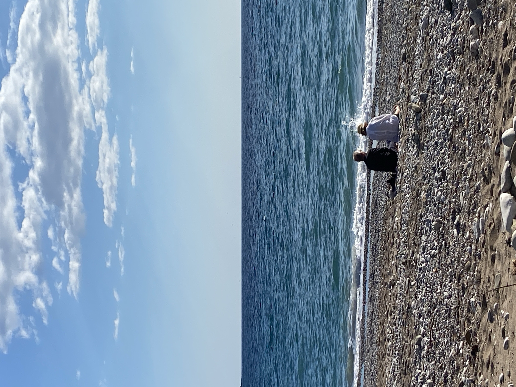 This image depicts a serene coastal scene in Bar, Montenegro, with a clear blue sky, calm sea, and a pebbly beach. The horizon line, stretching across the middle of the frame, separates the vast expanse of water from the sky.
In the foreground, two individuals are positioned near the water's edge on the pebble-covered shore. One person, wearing a light-colored top and a straw hat, is kneeling or sitting low to the ground, facing the sea. Slightly behind and to their left, another person, dressed in a dark top and dark pants, stands, also looking out towards the water. They appear to be observing the tranquil environment or possibly engaged in a leisurely activity on the beach.
The beach is characterized by numerous small, smooth, multi-colored pebbles and rocks. Gentle waves with white foam lap against the shore. The sea itself is a deep blue, showing subtle ripples on its surface under the sunlight. The sky is bright and clear, dotted with scattered, fluffy white clouds that add texture to the upper portion of the frame. The overall atmosphere suggests a peaceful and sunny day. No visible text is present in the image.