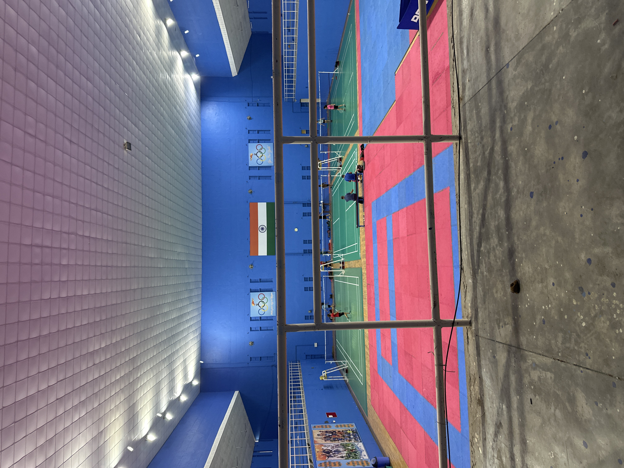 An indoor sports facility with blue walls and a white, paneled ceiling illuminated by rows of lights. The floor features a vibrant combination of pink and blue mats on the lower level, leading to green badminton courts with white nets on the upper level. Several people are visible, some playing badminton on the courts, while others are seated on the sidelines. Olympic rings and the Indian flag are displayed on the blue wall. A white railing in the foreground separates the viewer from the sports area. The scene appears to be at an indoor badminton and martial arts facility.