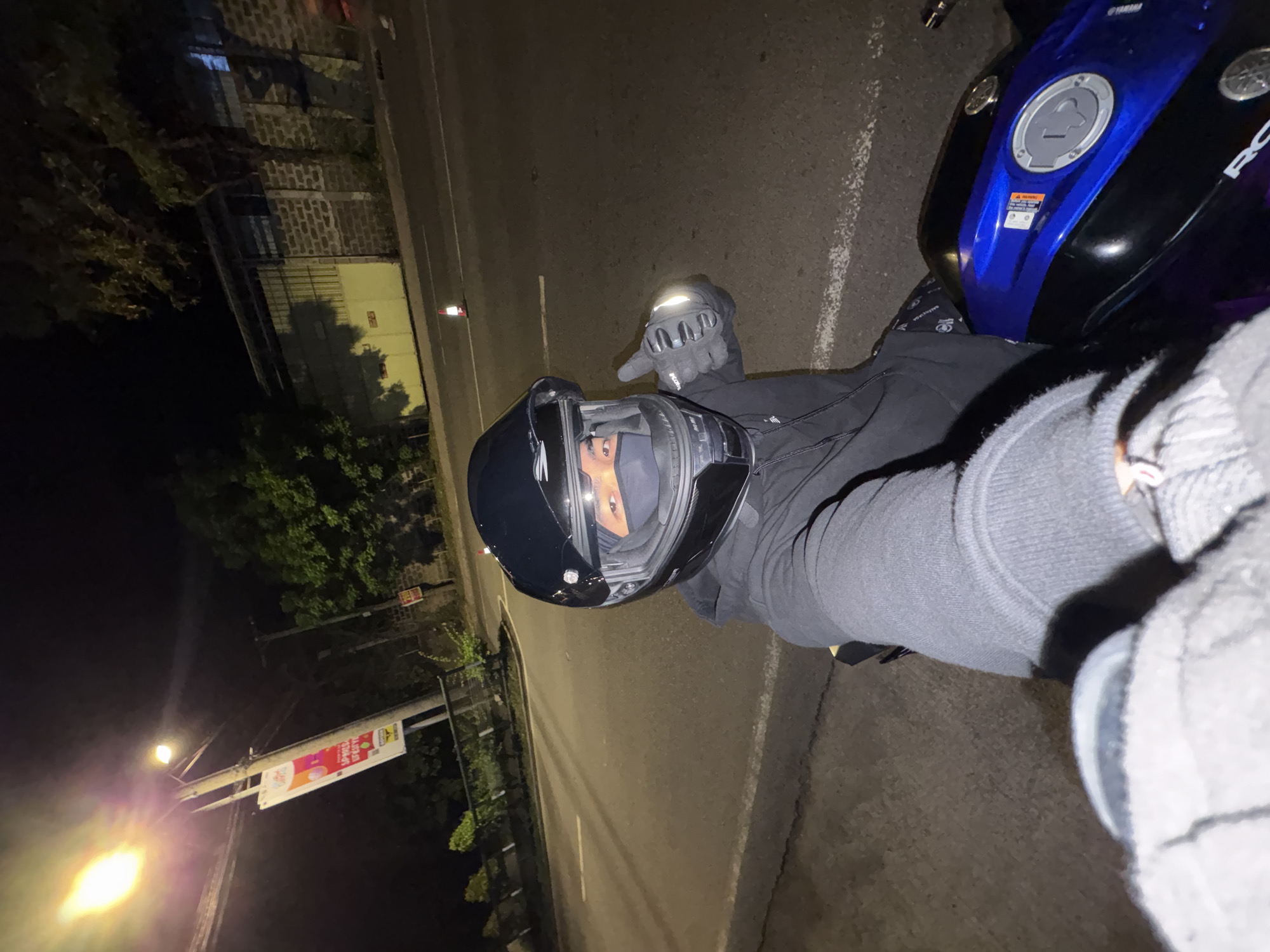 A first-person perspective selfie taken at night on a street in Davao City, Philippines, shows a person wearing a black motorcycle helmet, black face mask, black hoodie, and black gloves. The person is giving a thumbs-up gesture with their right hand, which is illuminated by a nearby light. The person is seated on a blue and black motorcycle. In the background, there is a street lamp, trees, a brick wall, and a yellow building with a corrugated metal roof. A traffic cone is visible on the road. A banner is hanging from a pole near the street lamp.