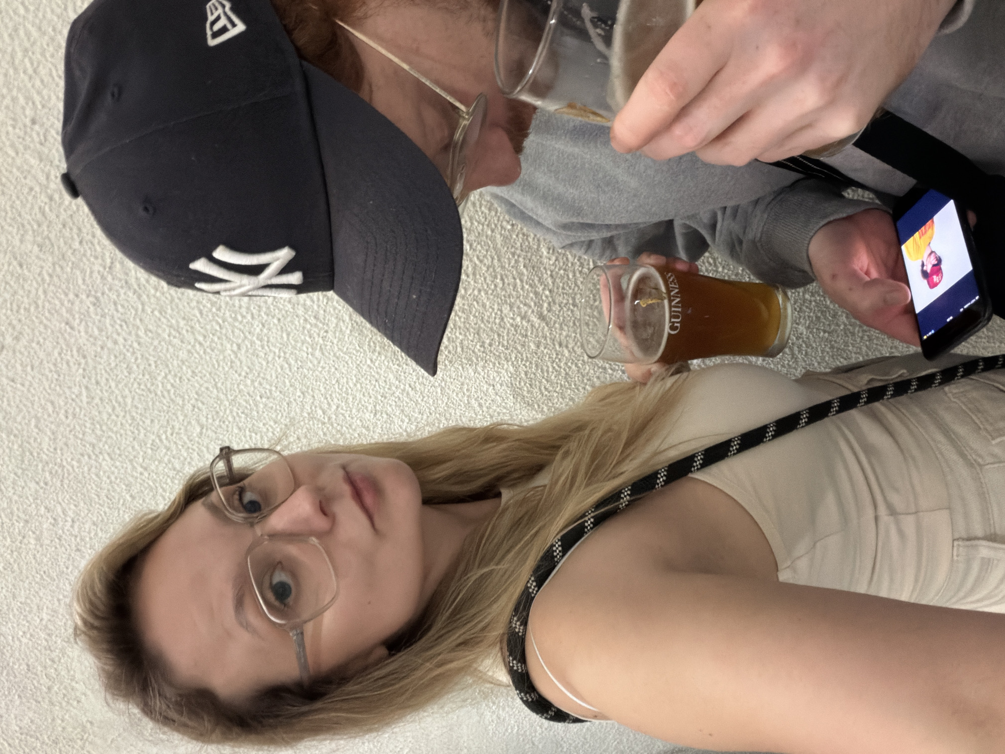 A group of three people are gathered indoors, likely in a pub setting. A young blonde woman, wearing glasses and a light-colored outfit, is looking directly at the camera. A man, wearing a dark blue baseball cap with the New York Yankees logo, is looking down at a beer glass in his hand. Another person is holding a smartphone displaying a picture. Glasses of beer are visible in the hands of the people. The location cannot be verified as being Tel-Aviv, Israel, from the image alone.