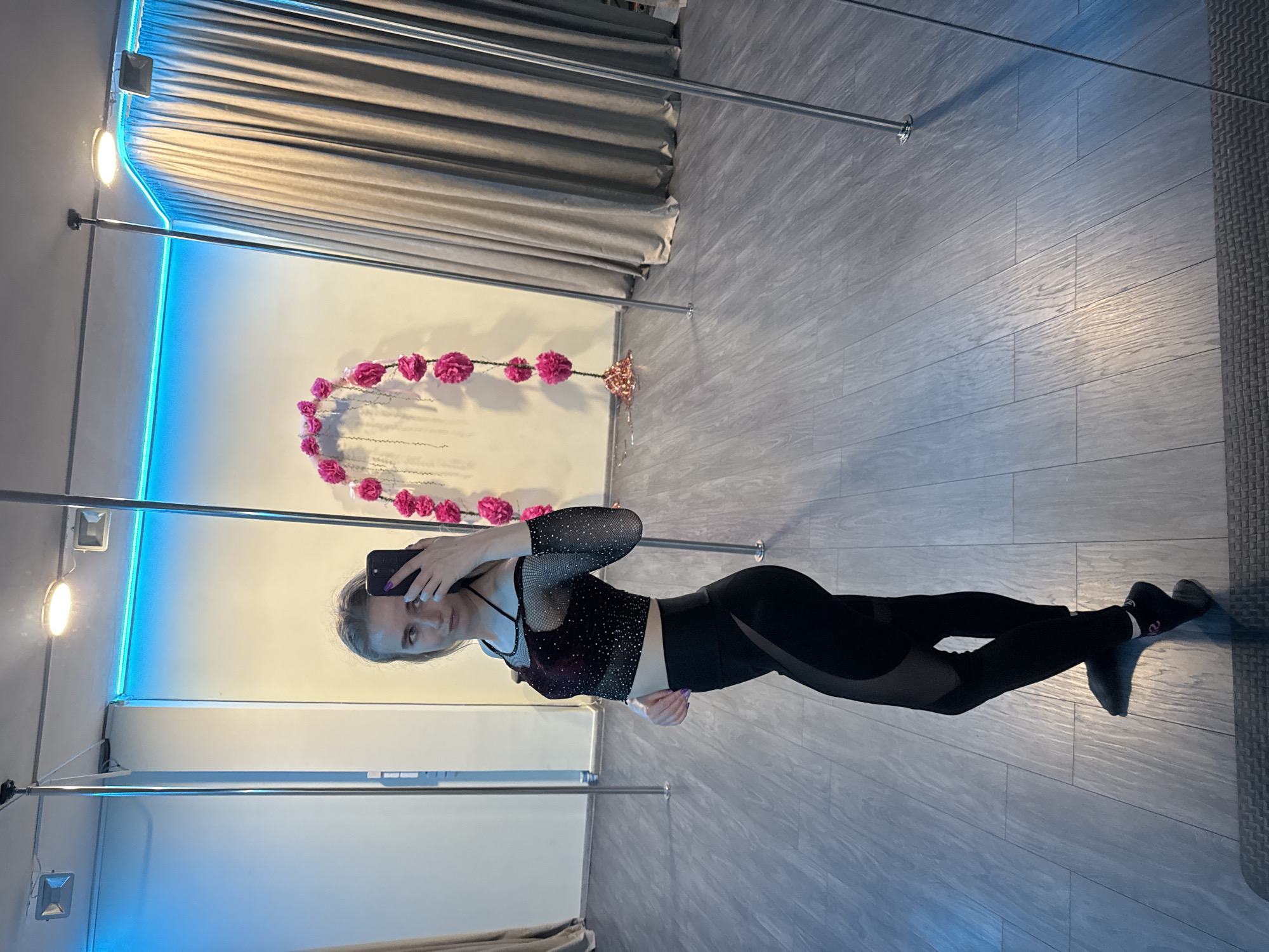 A woman is taking a selfie in a dance studio. She is wearing a black long-sleeved crop top with sparkles and black leggings with mesh panels. Her blond hair is pulled back. She is holding her phone in her left hand, taking the picture. The background features a white wall with a garland of pink flowers, a blue neon light strip, and silver curtain rods. The floor is gray wood-look vinyl.