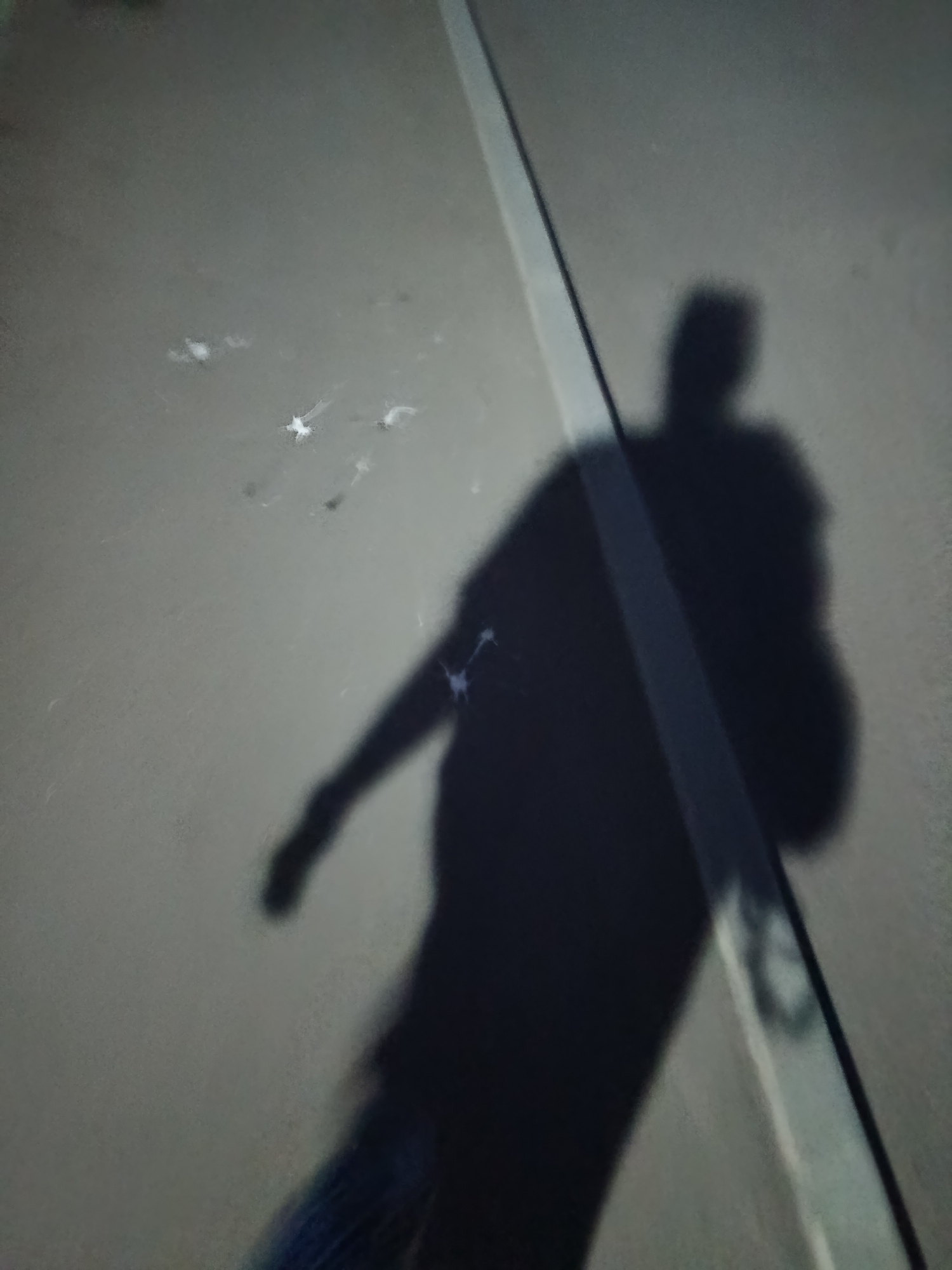 An underexposed, medium-shot, low-angle photograph captures the shadow of a person walking on a paved surface, possibly a road or sidewalk, marked by a white line. The shadow is dark and amorphous, with a discernible head, torso, and limbs. The person appears to be carrying something on their right side, creating a bulky mass in the shadow. Scattered on the ground are small, bright white specks, possibly debris or reflections. The environment appears to be outdoors in a city. The shadow's orientation suggests the sun is to the left and slightly behind the photographer, casting the shadow forward. The overall scene is gritty and urban.