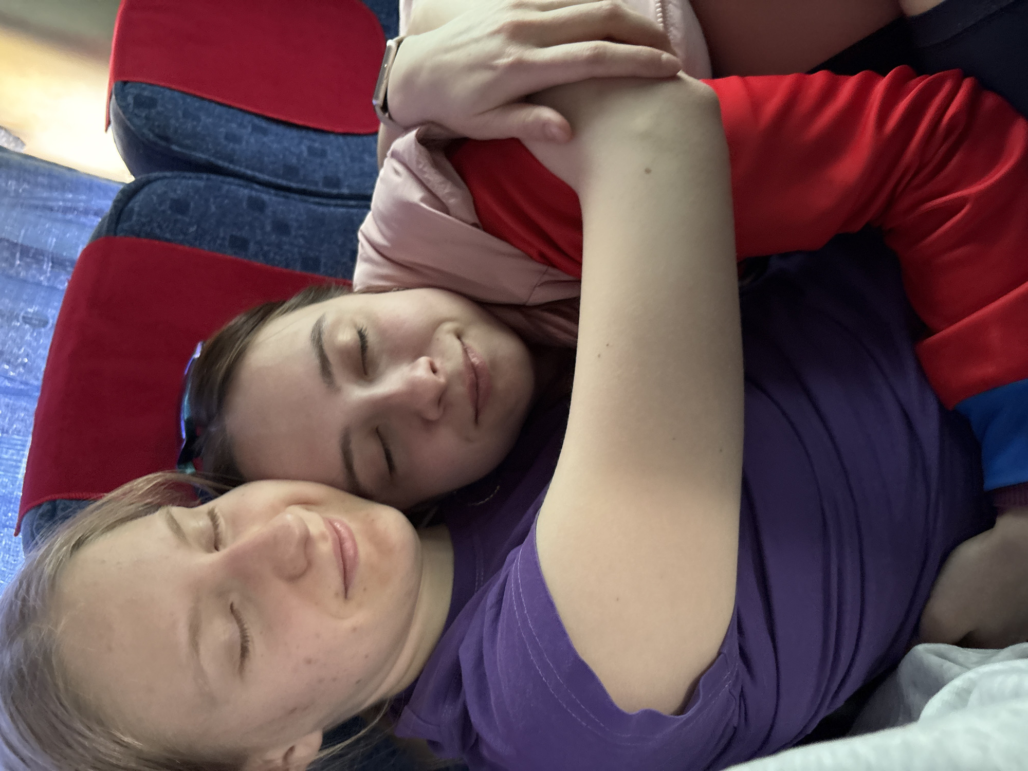 Two young women are sleeping on a bus seat. The woman on the bottom is wearing a purple t-shirt and her eyes are closed. The other woman is lying on top of her, wearing a red long-sleeved shirt and a pink jacket. Her eyes are also closed. The seat is upholstered in a red and blue patterned fabric. The location is Verkhnyaya Dobryanka, Russia, indicated by the presence of a bus often used for travel within that region.