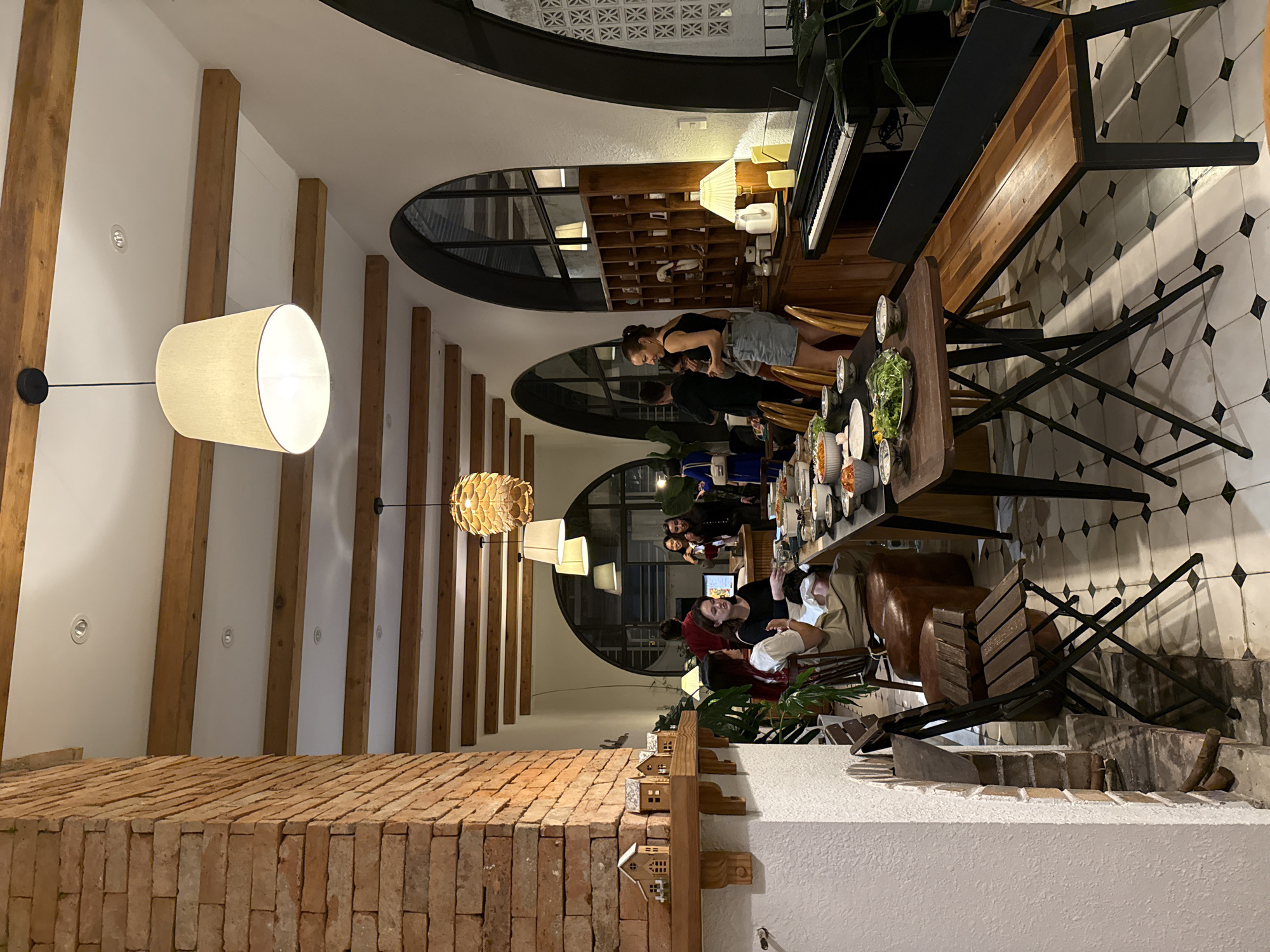 The image depicts an indoor gathering at what appears to be a restaurant or home in Lam Vien - Da Lat, Vietnam. The setting is rustic and cozy, with exposed wooden beams on the ceiling and a prominent brick pillar on the left. A long dining table, set with numerous bowls of food and plates of greens, is the central focus. Several people are seated around the table, engaged in conversation or eating. Others are standing and interacting, some near a counter with a computer monitor. In the background, there are arched windows and a wooden cabinet filled with decorative items. A black digital piano is visible to the right of the cabinet. The flooring is tiled with a black and white checkered pattern. The overall atmosphere suggests a social event or meal.