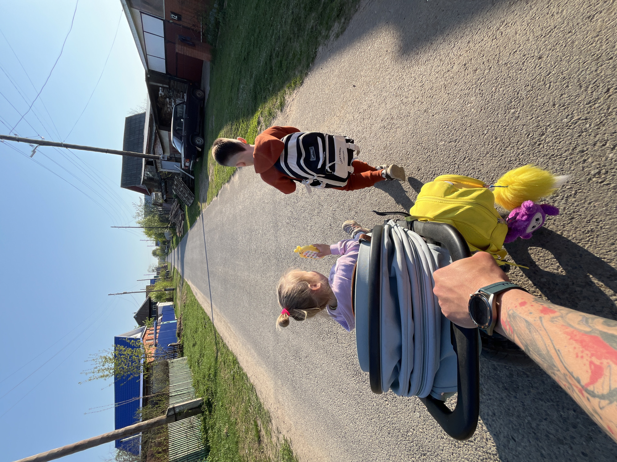 A ground-level, medium, eye-level shot captures a person pushing a stroller down an asphalt road in what appears to be a residential area in the city of Chaikovsky, Russia. The camera is presumably held by the person pushing the stroller, as an arm with tattoos and a wristwatch is visible in the foreground, gripping the stroller handle.
A young girl in a light purple jacket is seated in the stroller. She holds a yellow toy in her hand. Attached to the stroller is a bright yellow backpack with a fluffy yellow tail and a purple stuffed animal dangling from it.
A young boy in an orange tracksuit with a black and white backpack walks slightly ahead and to the right of the stroller. He walks away from the camera.
The asphalt road stretches into the distance, flanked by grassy areas and houses. On the left, a blue-roofed house and a green fence are visible. Power lines extend across the sky. The day appears sunny.