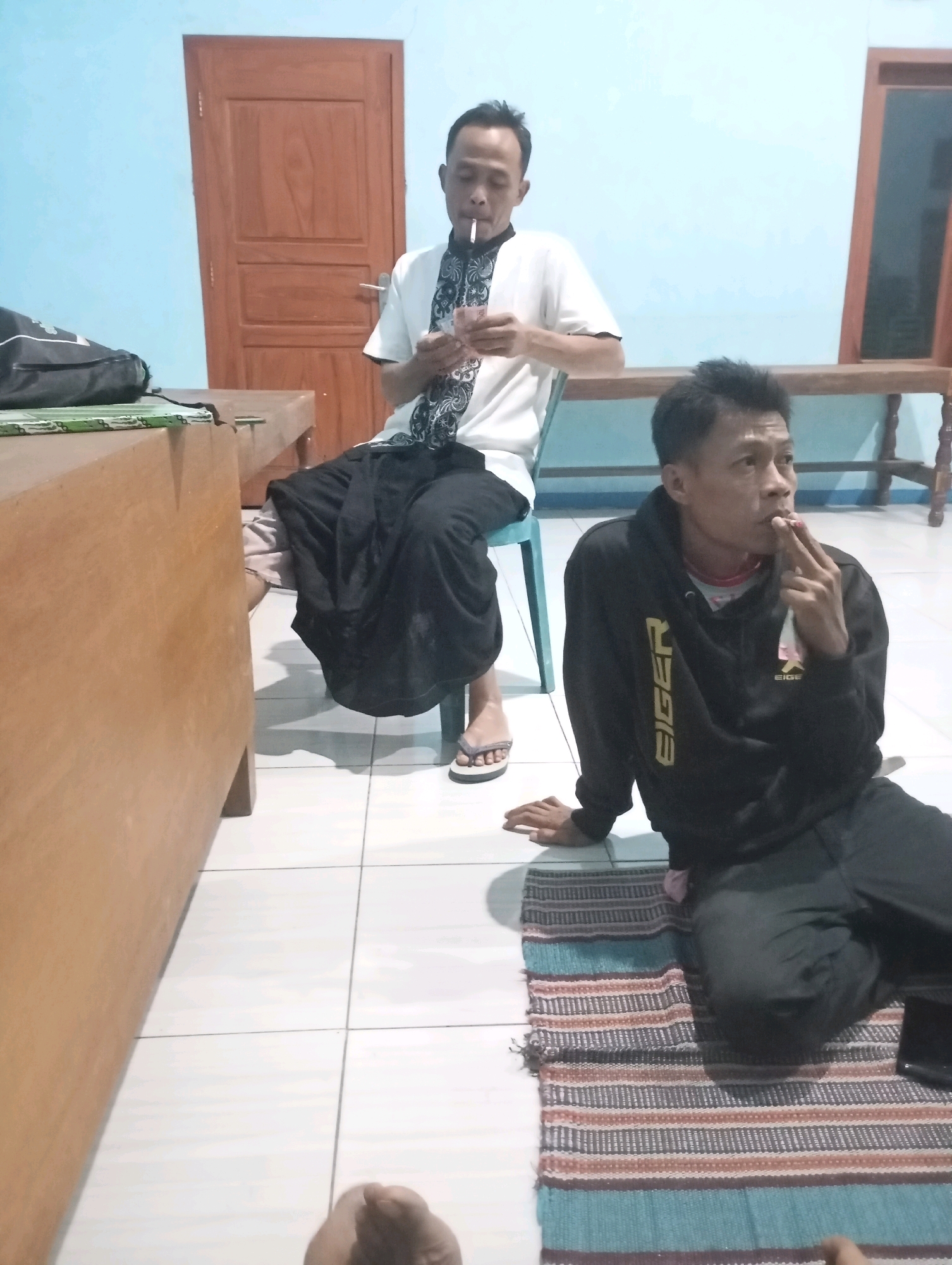 Two men are in a room, likely in Ketepung, Indonesia. The man in the background sits on a chair wearing a white shirt with a black and white patterned collar, and black pants. He is holding paper money and a cigarette in his mouth. The man in the foreground sits on a colorful rug on the floor, wearing a black hoodie with "EIGER" in gold lettering and black pants. He is smoking a cigarette and holding it with his fingers. The room has light blue walls and a wooden door in the background. A wooden table and chair are also visible, along with a bag on the table. The floor is tiled.