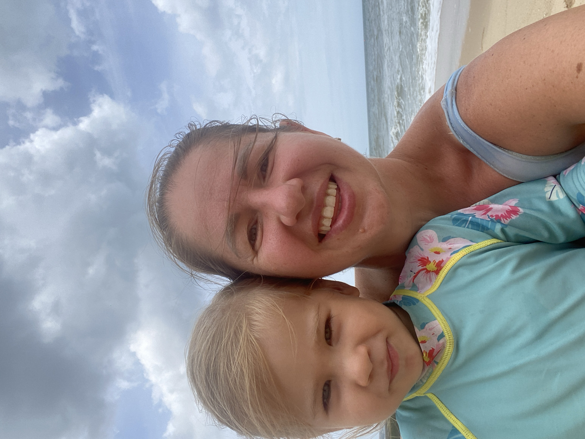 A woman and a young child are taking a selfie on a beach. The woman is smiling, and the child is looking at the camera. They are both wearing beachwear. The beach is sandy, and the ocean is visible in the background. The sky is cloudy. The location is the city of Mui Ne, Vietnam.