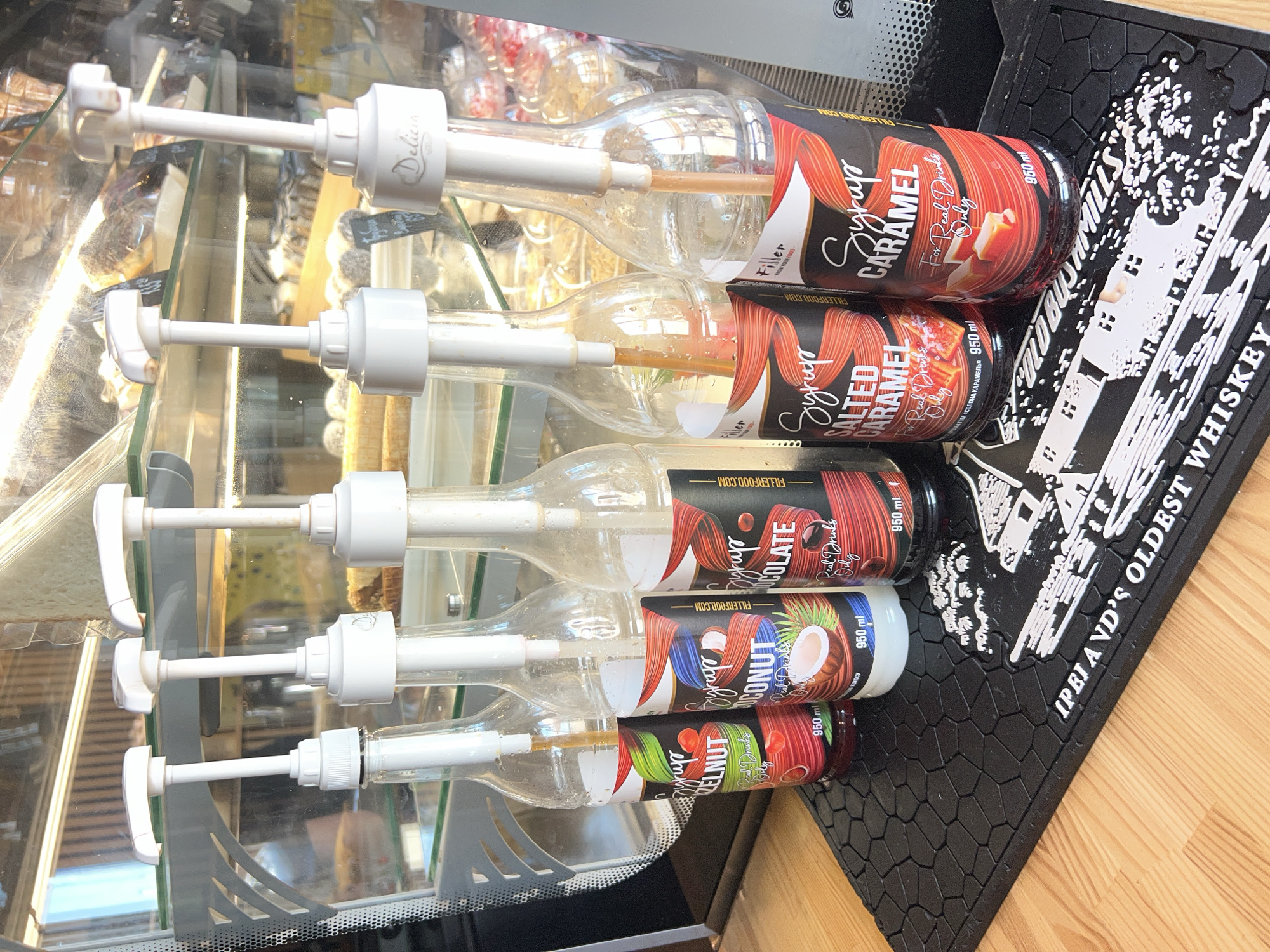 The image shows a close-up of five bottles of syrup with white pump dispensers, arranged in a row on a black mat with a textured pattern. The bottles are clear and contain various colored syrups. The labels on the bottles are visible, with some of the text readable. From left to right, the visible labels indicate "Syrup HAZELNUT," "Syrup COCONUT," "Syrup CHOCOLATE," "Syrup SALTED CARAMEL," and "Syrup CARAMEL." The syrups appear to be for use in drinks, as suggested by the text "For Real Drinks Only" on some of the labels. The bottles are in a cafe or bar setting, with a glass display case in the background containing baked goods and other items. The mat on which the bottles sit has a raised design that appears to depict buildings and the words "IPEIAND'S OLDEST WHISKEY." The lighting suggests an indoor setting, possibly with natural light coming from a window or overhead. There are no people visible in the image. The location in Fontanka, Ukraine is not discernible from the image itself.