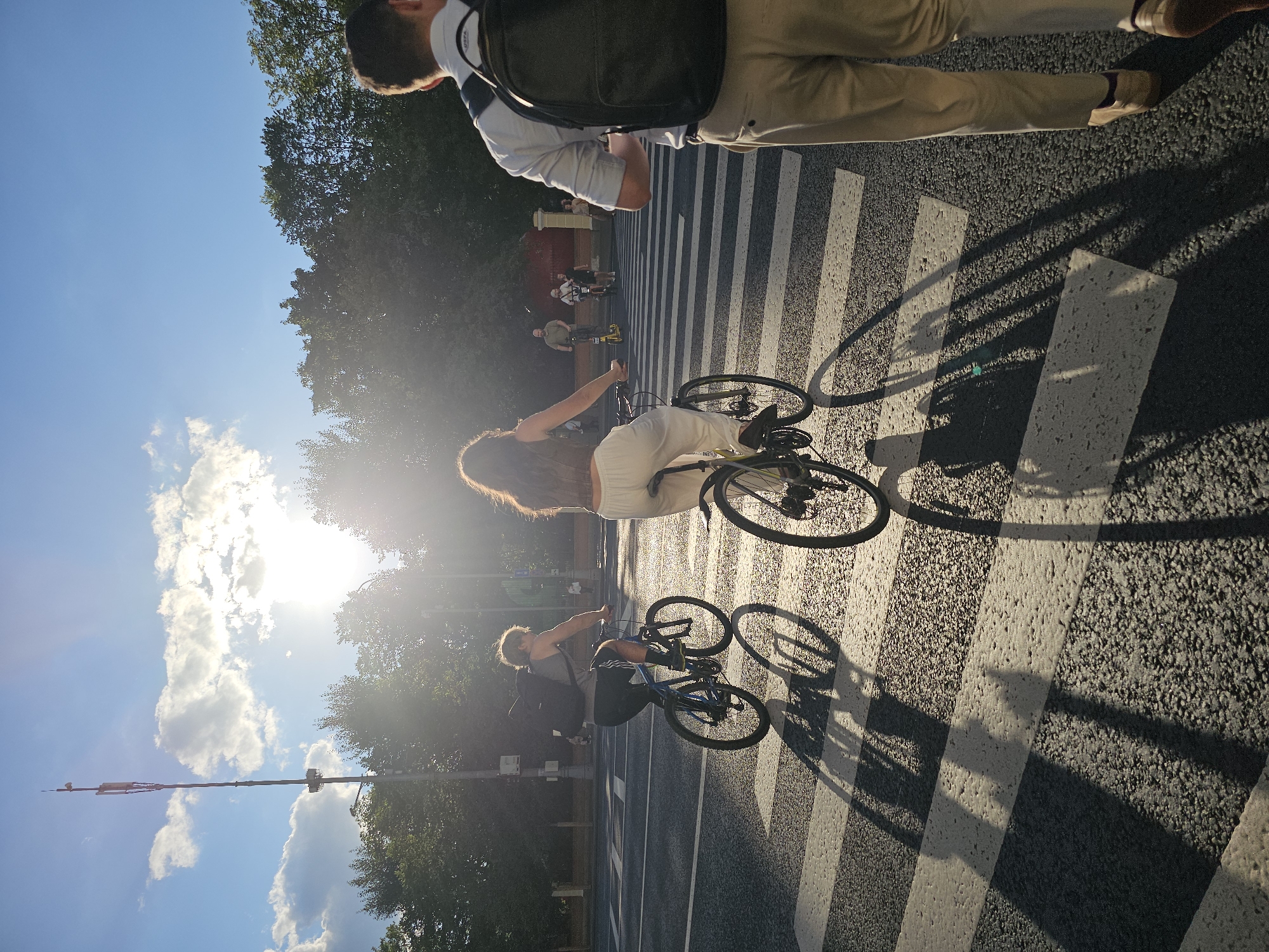In Moscow, Russia, a street scene unfolds at a pedestrian crossing under a bright, sunlit sky. The sun is low in the sky, casting long shadows across the zebra stripes of the crosswalk. Two individuals are riding bicycles across the crossing, their shadows stretching behind them. The person in the foreground on the right is walking away from the camera, wearing a light-colored shirt, khaki pants, and a black backpack. In the background, several other people are visible, some on what appear to be electric scooters, and trees line the street, creating a backdrop of greenery. A tall communication tower stands prominently against the blue sky, with scattered clouds. The overall impression is a snapshot of urban life during a sunny day.