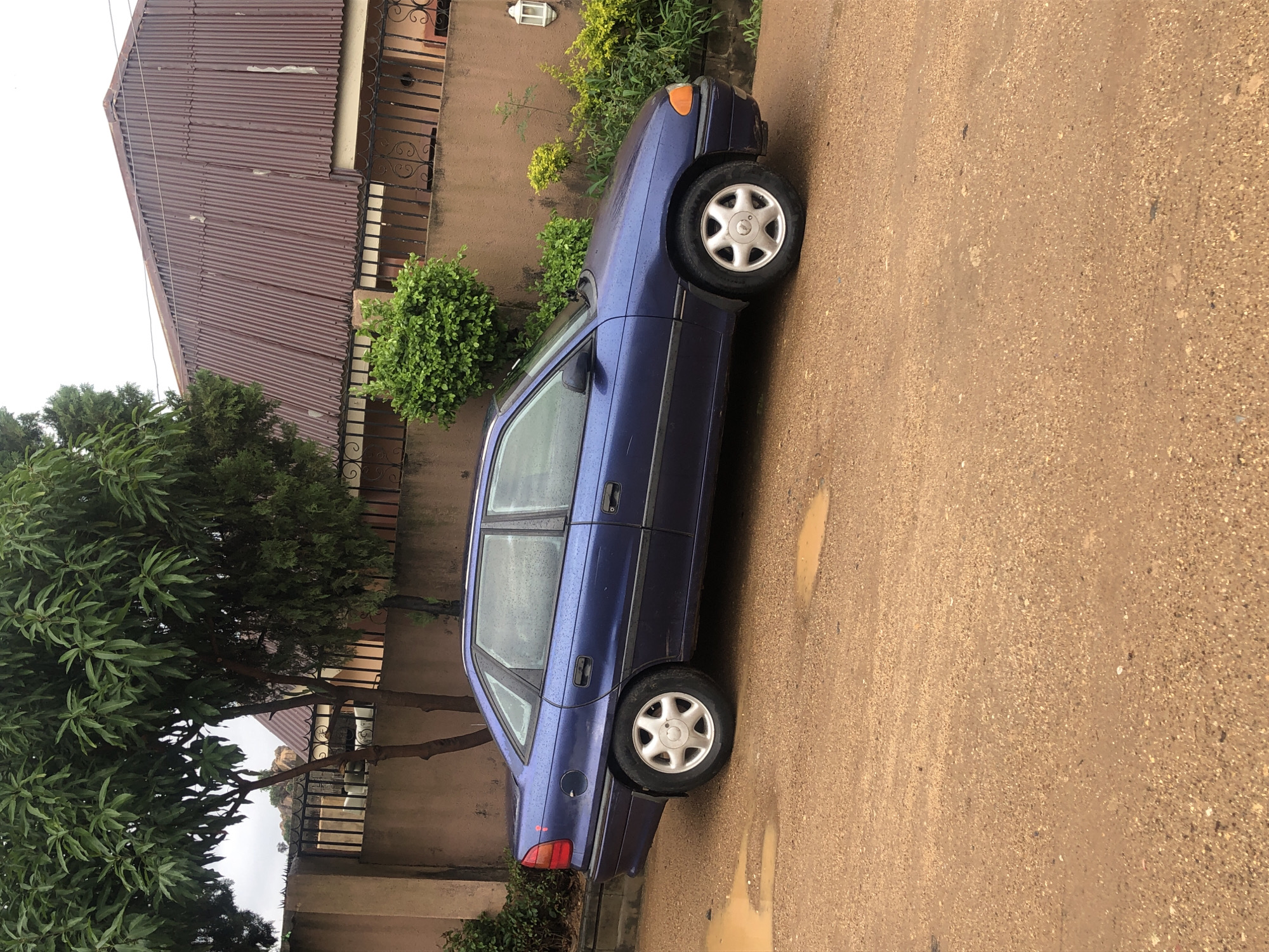 A dark blue sedan is parked on a wet, dirt road. The car is viewed from the side, with its front facing to the right of the frame. The road is covered in puddles, suggesting it has recently rained. In the background, there is a light brown wall topped with a black metal fence. Behind the fence, there are trees and a house with a corrugated metal roof. The sky is overcast.