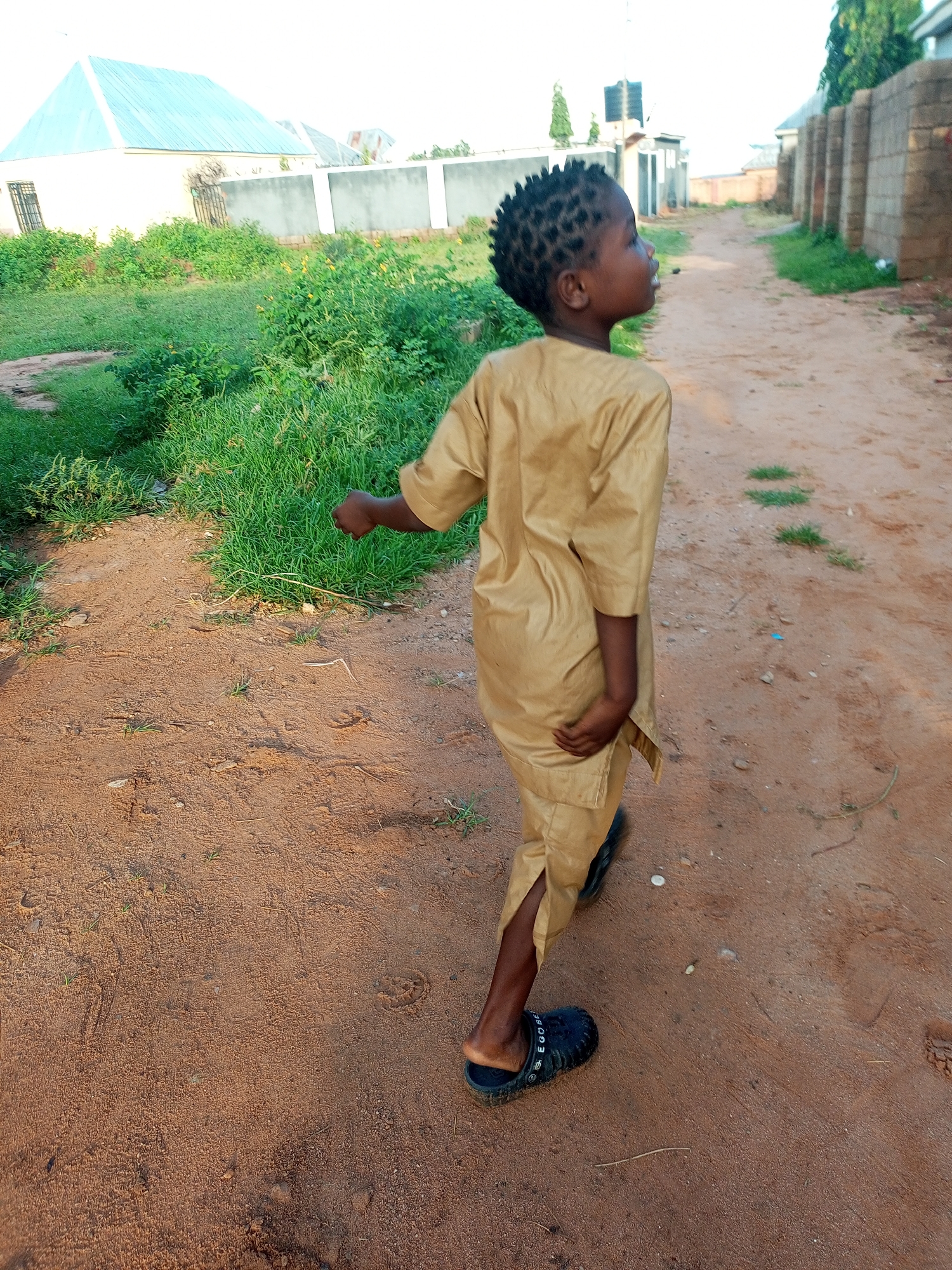 A young boy is walking on a dirt road in Tempure, Nigeria. He is wearing a tan colored traditional outfit and black slides. The road is lined with greenery on the left and a brick wall on the right. Buildings can be seen in the background. The sky is overcast.