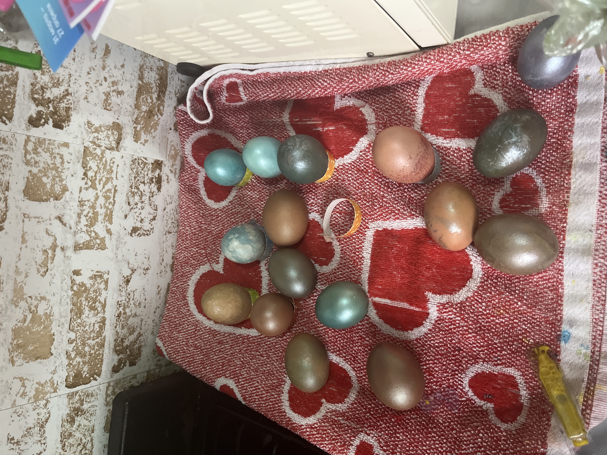 The image shows a collection of approximately twenty painted Easter eggs scattered on a red and white patterned towel. The eggs are various shades of brown, blue, and metallic colors. Small pieces of yellow tape or stickers are visible near some of the eggs. The towel is placed on a surface next to a tiled wall and part of a white appliance, possibly an oven or microwave. The scene appears to be indoors. There is no indication that the image was taken in the city of Oryol-Izumrud, Russia.