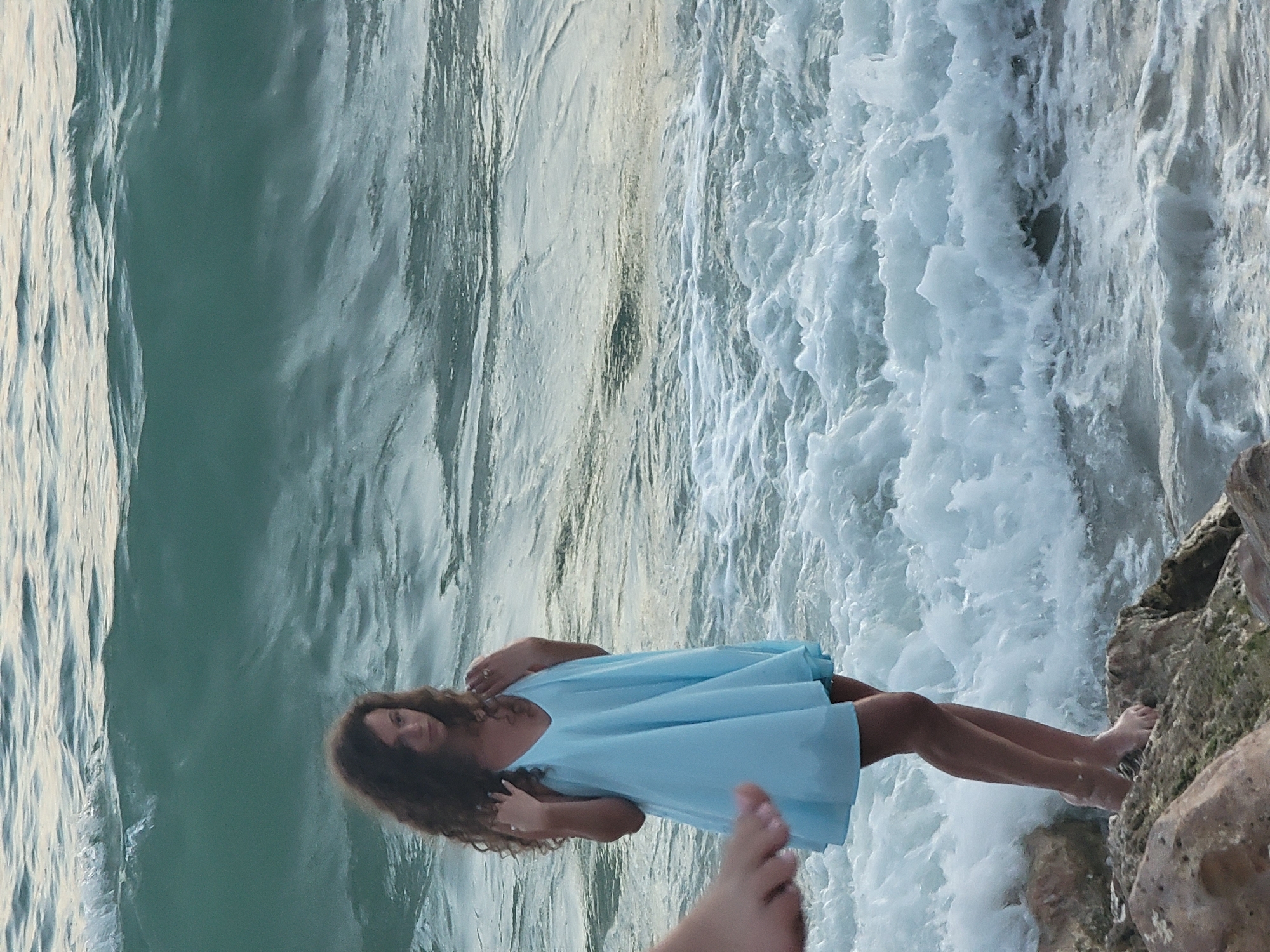 A young woman with curly brown hair stands on rocks at the edge of the ocean, wearing a light blue sundress. The waves are breaking around her, with white foam and turquoise water. Her feet are bare and wet, and she is wearing rings on her fingers. In the foreground, a person's foot is visible, also at the edge of the frame. The scene appears to be a beach in Quarteira, Portugal, likely during the day. The lighting suggests it may be late afternoon or early morning. The overall atmosphere is serene and natural.