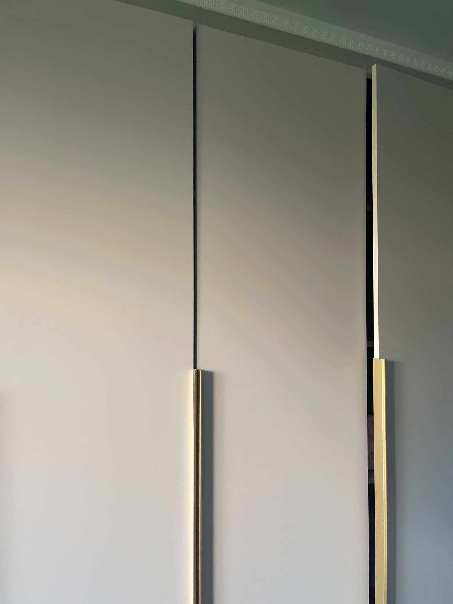 The image shows a close-up of a section of a wardrobe with three doors. The doors are a light grey color and are flush with each other, with thin vertical black gaps between them. Each door has a vertical, rectangular gold handle positioned slightly below the center. The wardrobe is positioned against a wall which appears to have a decorative border along the top. The lighting suggests the wardrobe is indoors, possibly in a residential setting. The location in Tallinn, Estonia, is not discernible from the image itself.