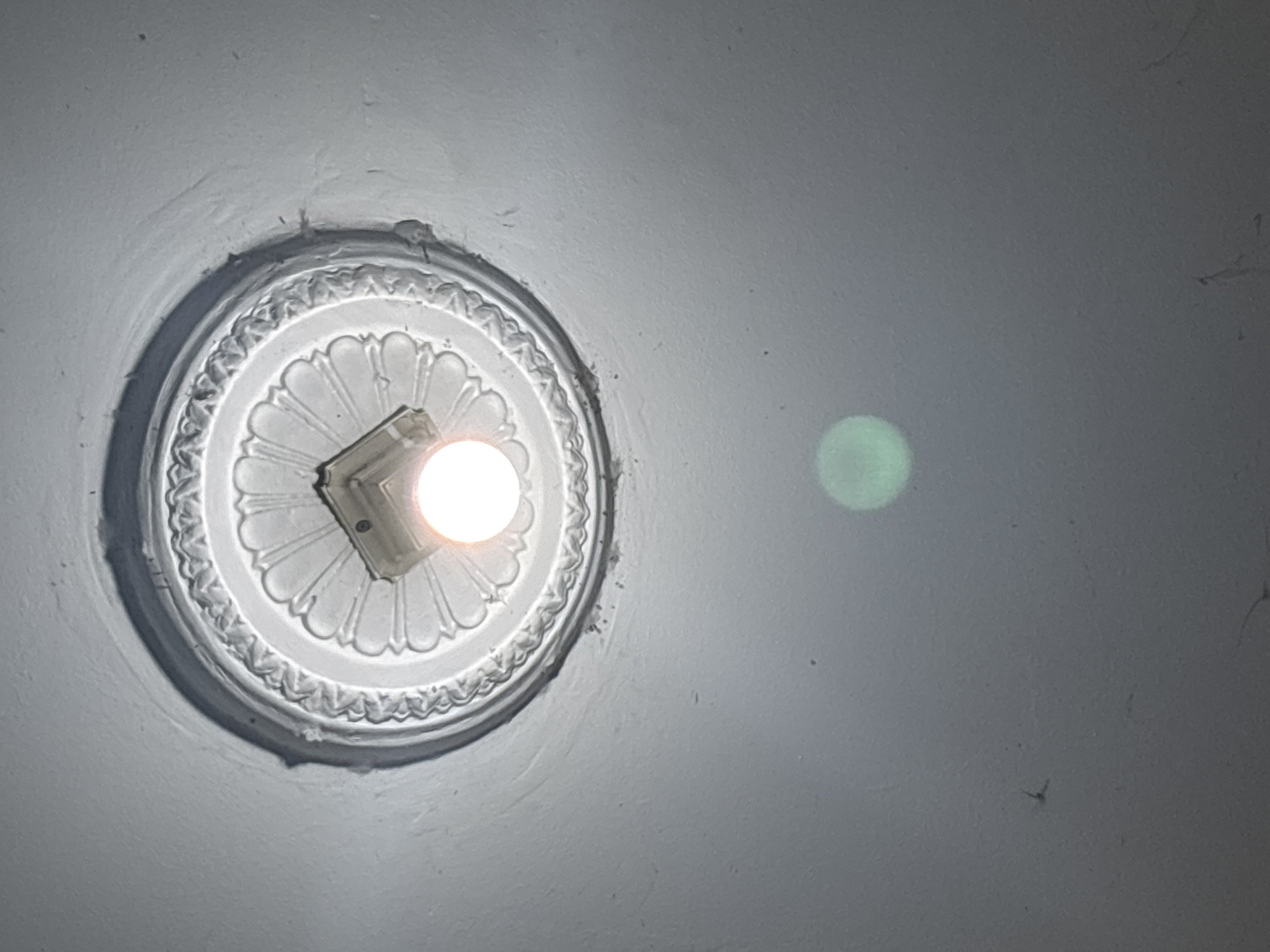 A low-angle shot of a white ceiling with a decorative medallion and a lightbulb. The medallion is circular and features a raised, ornate pattern around its edge and a fluted design radiating from the center. A square light fixture with a visible lightbulb is centrally mounted within the medallion. The lightbulb is illuminated and emits a warm glow. Below the medallion, a soft, circular green light artifact is visible on the ceiling, possibly a lens flare or reflection. The ceiling itself appears to have minor imperfections and some faint spiderwebs. The image is taken from below, looking upwards towards the ceiling.