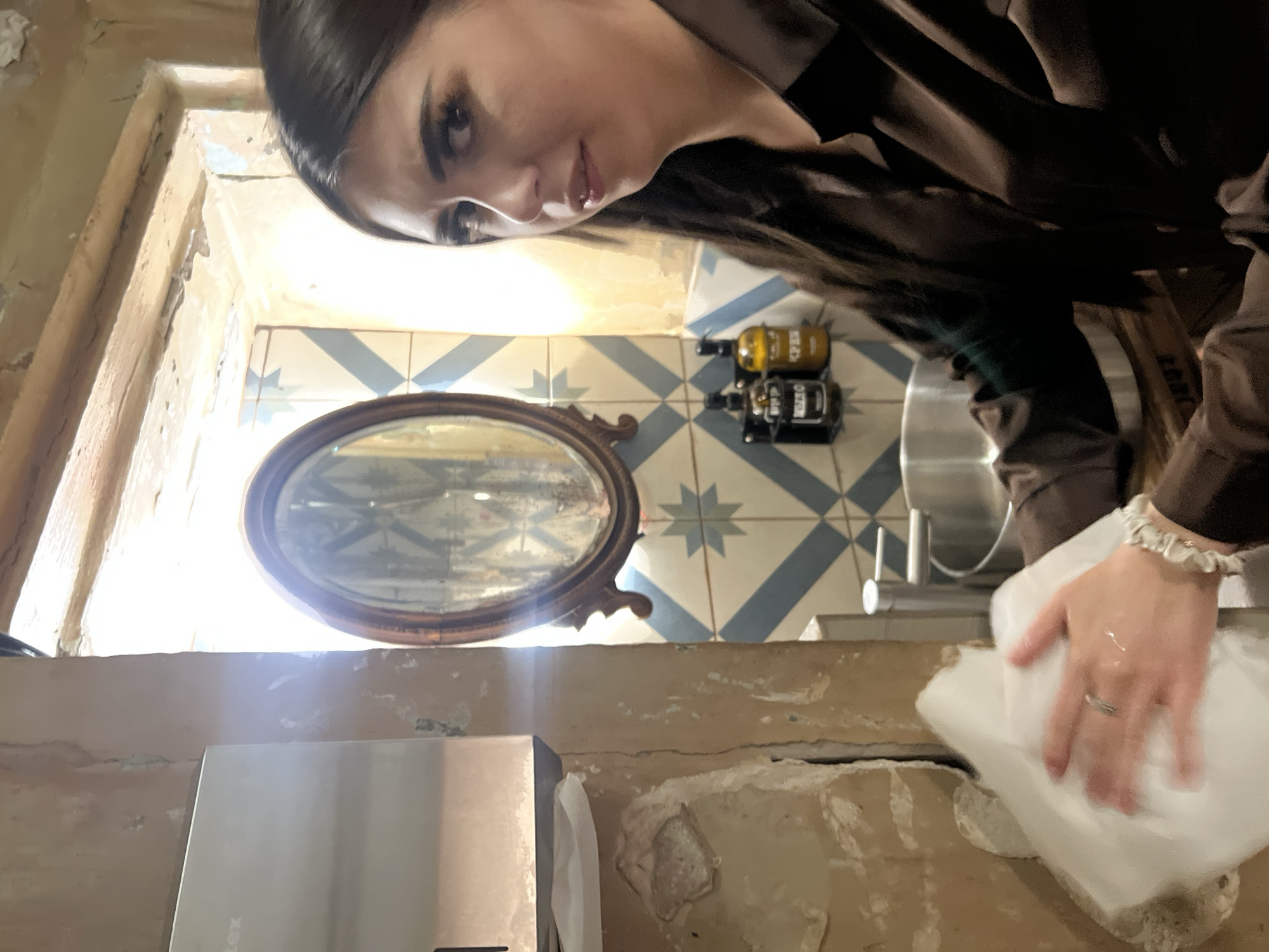 The image depicts a woman with dark hair and makeup, wearing a brown shirt, standing in what appears to be a dimly lit room with a rustic aesthetic. She is holding a white paper towel and her hand is in motion, suggesting she is in the process of cleaning or wiping something. To her left, a dispenser is mounted on a textured, peeling wall. Behind her, a sink is visible, along with two bottles of liquid soap or lotion on the wall. An oval mirror with a wooden frame hangs on a wall decorated with blue and white patterned tiles. The overall environment suggests a restroom or a wash area in a restaurant or similar establishment. The lighting is dim, with a strong source of light coming from behind the mirror, creating a glare and obscuring some details. There is no discernible connection to Nizhny Novgorod, Russia, in the image.