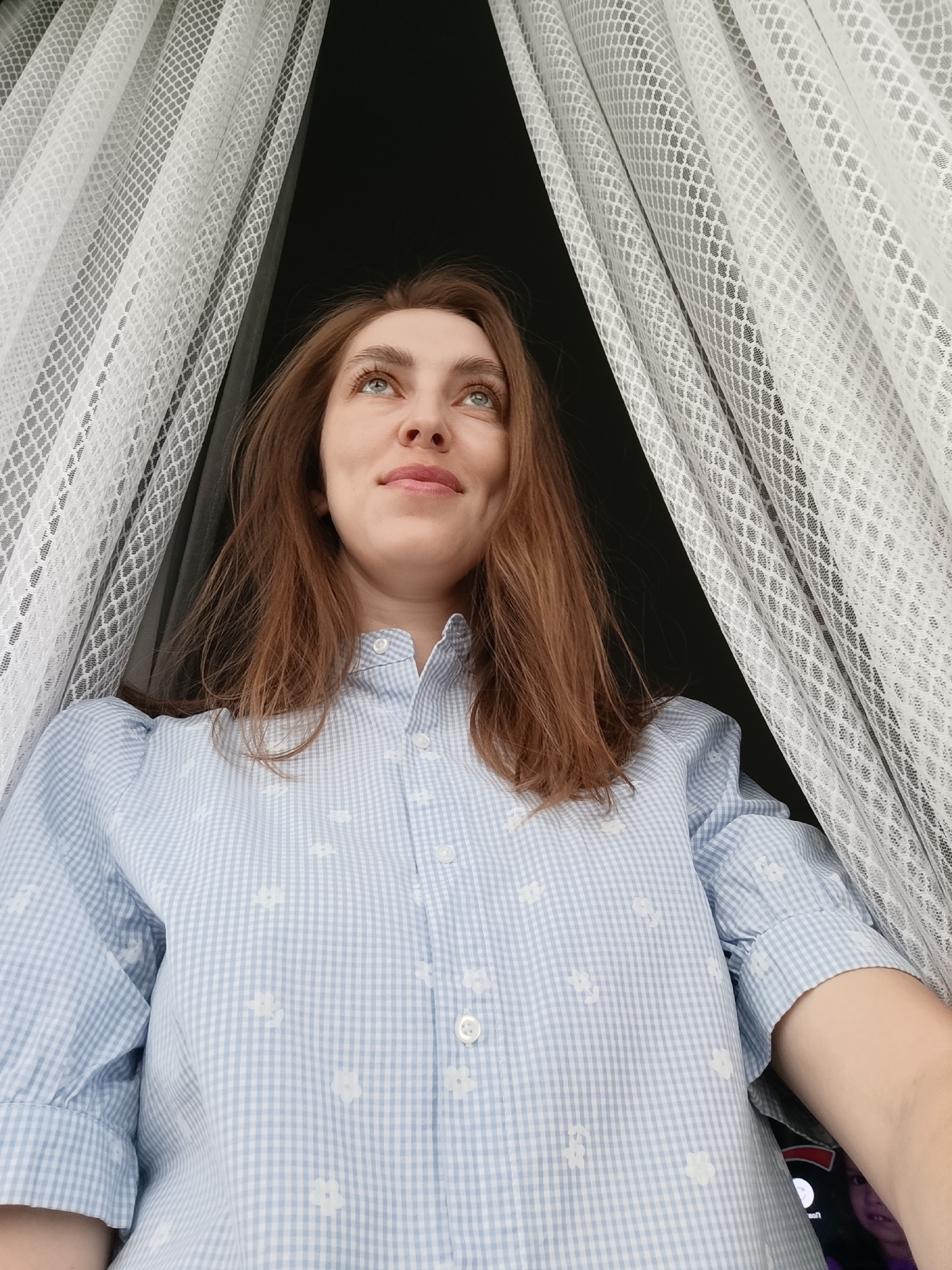 A low-angle medium shot depicts a fair-skinned woman with reddish-brown hair, dressed in a light blue checkered shirt with white floral patterns, looking upwards with a slight smile. Her gaze is directed towards the upper left corner of the frame. She is positioned between two panels of white, semi-transparent net curtains. The background behind the curtains appears to be dark, possibly a window at night or a dimly lit interior. The location is Chaikovsky, Russia.