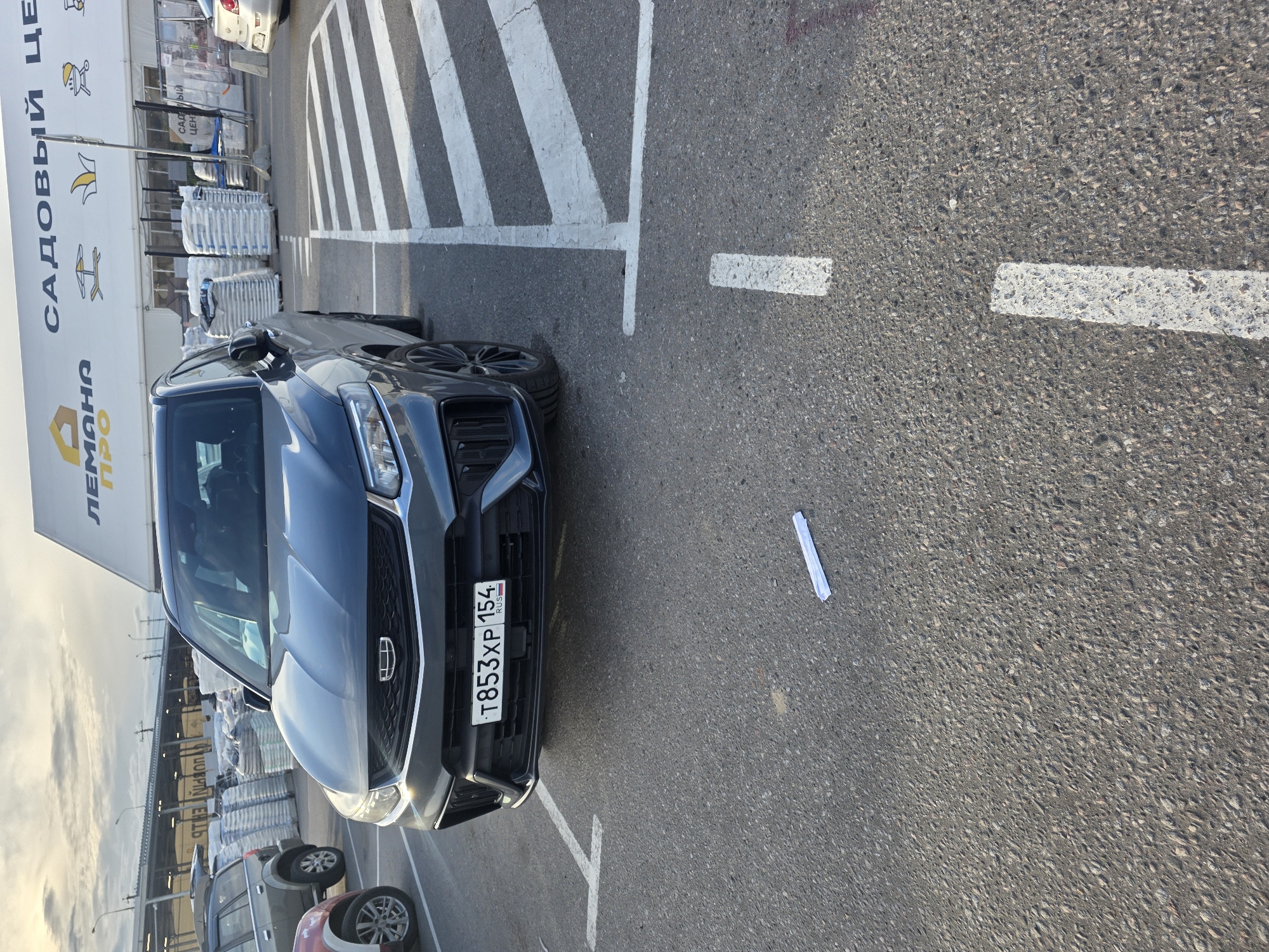 This media file shows a dark grey Geely car parked in a lot with a large building in the background. The building has a prominent sign with Russian text that reads "САДОВЫЙ ЦЕНТР" and "ЛЕМАНА ПРО". The car has a license plate that reads "T853XP 154". White painted lines on the asphalt indicate parking spaces. There are stacks of bagged goods behind the car, possibly related to the garden center. The sky suggests it is either early morning or late afternoon, with soft lighting. The scene appears to be a commercial area, likely a garden supply store. There are no people visible in the image.