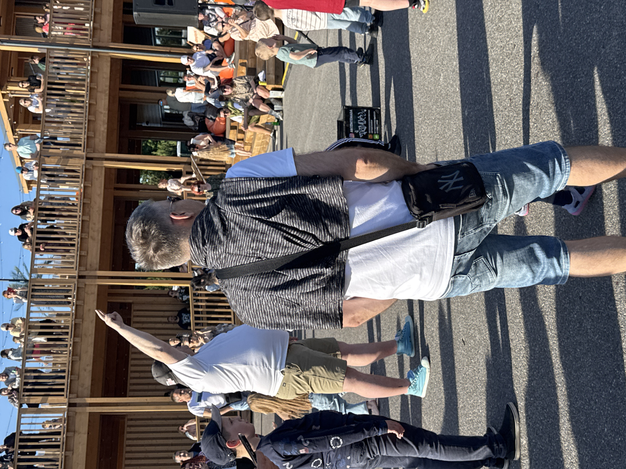 The image shows a group of people gathered outdoors in what appears to be a public event or gathering. The setting is a multi-story wooden structure with balconies, possibly a building with outdoor seating or a market. The people are standing and observing something happening in front of them. Some individuals are looking upwards, suggesting they are watching a performance or an announcement.
One man in the foreground is wearing a striped vest over a white t-shirt and denim shorts. He has a black pouch or bag slung across his chest. Another man in the middle ground is wearing a white t-shirt and khaki shorts, with his right arm raised and pointing. A young boy in the background is wearing a green t-shirt and dark pants, standing with his hands behind his back.
The lighting suggests it is daytime, with strong shadows indicating a sunny day. The presence of multiple people suggests a social event. The wooden architecture might be typical of a certain region or style, and the mention of Tallinn, Estonia, provides a specific geographical context. No specific event is identifiable, but the gathering appears to be casual and community-oriented. There is some visible text on a black box in the background which is illegible.