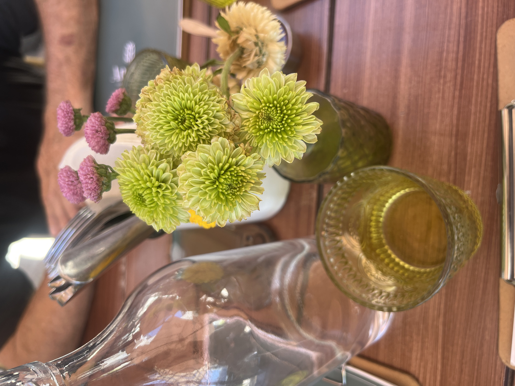 A close-up, overhead shot shows a still life arrangement on a brown wooden surface. A clear glass bottle lies horizontally, partially out of frame at the bottom. Next to it is a green-tinted glass with a textured exterior. A vase holds green chrysanthemum-like flowers and pink globe amaranth flowers. Cutlery, including a fork, sits alongside the flowers. The arrangement appears on a table, possibly in a restaurant. It is difficult to confirm the location as San Bernardino Tlaxcalancingo, Mexico, from the image alone.