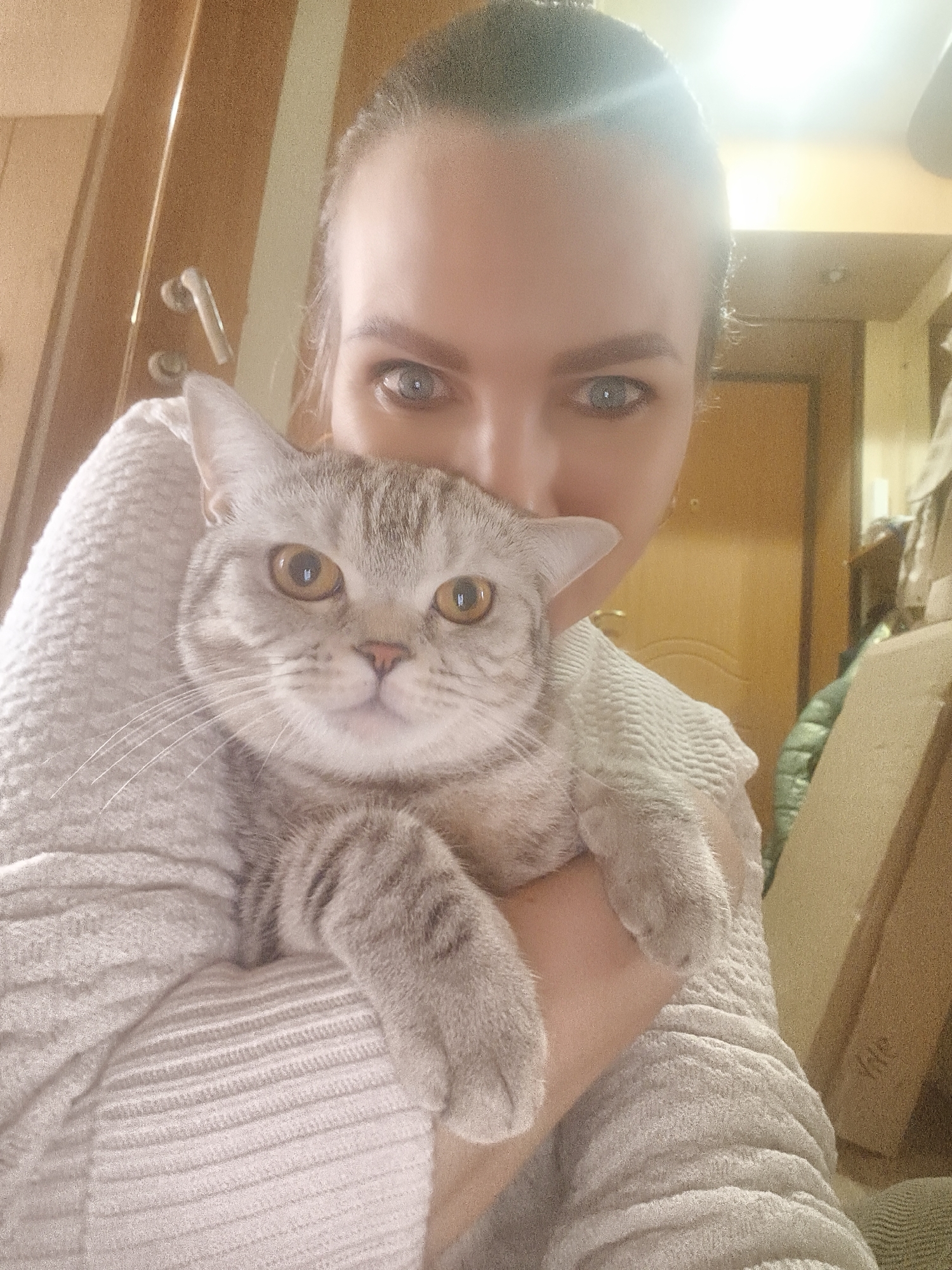 This close-up portrait features a woman holding a cat. The woman's face is largely obscured by the cat, with only her upper face visible. She has striking blue-grey eyes and her hair is pulled back. She is wearing a light-colored, possibly cream or very light grey, sweater with a distinct ribbed and textured pattern.
The cat, positioned centrally in the foreground, is a light grey tabby with subtle stripes and short fur, characteristic of a British Shorthair breed. It has prominent, round amber-orange eyes and a small pink nose, and appears calm and comfortable in the woman's arms. Its front paws rest on the woman's arm, which is clad in the textured sweater. Both the woman and the cat are looking directly at the camera.
The setting is indoors, with a blurred background showing warm-toned wooden elements, likely a door or furniture panels. A silver-colored door handle is visible on the left side of the frame. A bright light source, possibly overhead or from a window, creates a noticeable lens flare at the top center of the image, contributing to the overall warm lighting of the scene. No visible text is present in the image.