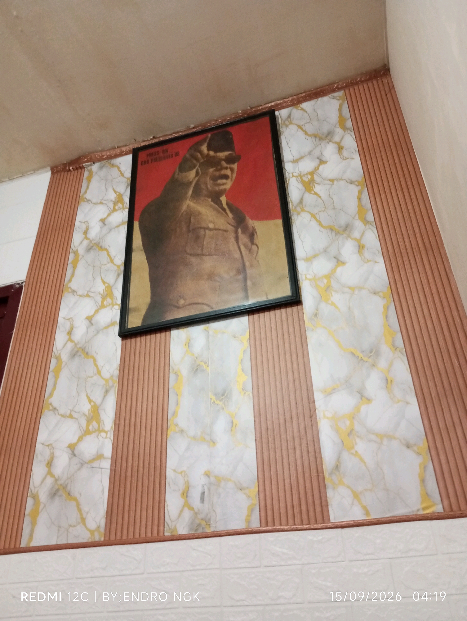 An indoor scene features a framed portrait of a man in military uniform, possibly a historical figure, displayed prominently on a wall. The wall is decorated with alternating vertical panels of marble-patterned wallpaper with gold veining and a textured, wood-like material in a reddish-brown hue. A white brick-patterned wallpaper is visible at the bottom of the frame. The ceiling appears to be a standard white material. The timestamp "15/09/2026 04:19" is present in the lower right corner, along with "REDMI 12C | BY:ENDRO NGK" in the lower left, indicating the image was captured with a Redmi 12C phone by Endro Ngk on September 15, 2026, at 4:19 AM. The context of Ringinanom, Indonesia, suggests a potential location within a residence or public space in that area.