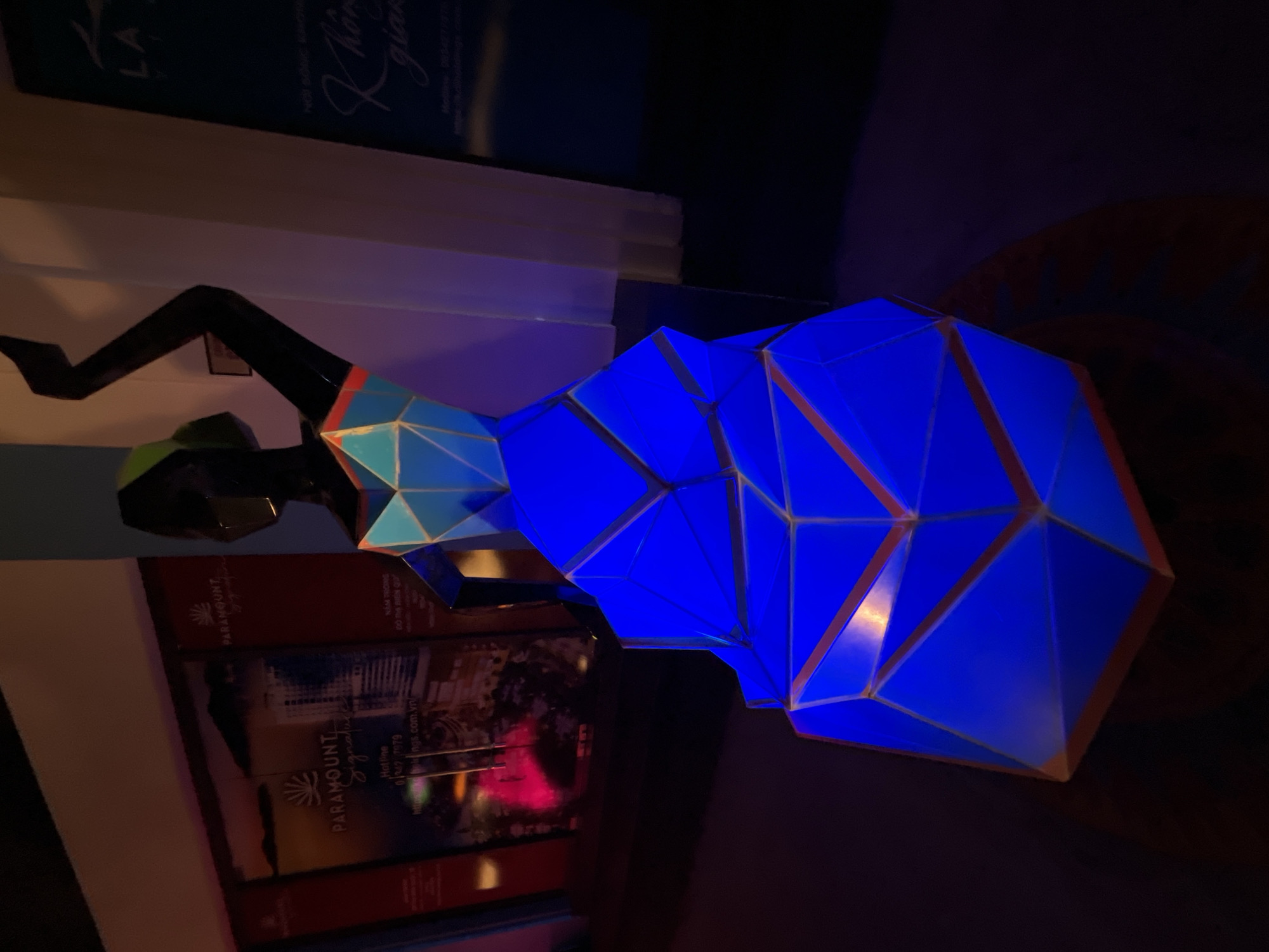 The image depicts a modern sculpture of a woman's figure, illuminated with bright blue light, and composed of geometric facets. The sculpture has a black base, suggesting legs and torso, with a polygonal head and dress. It sits on a shelf, positioned next to a stack of promotional items. The promotional items are labeled "PARAMOUNT Signature" and contain details about Bac Nha Trang Ward, Vietnam. The area is referred to as an urban waterfront. The light conditions of the room are subdued.