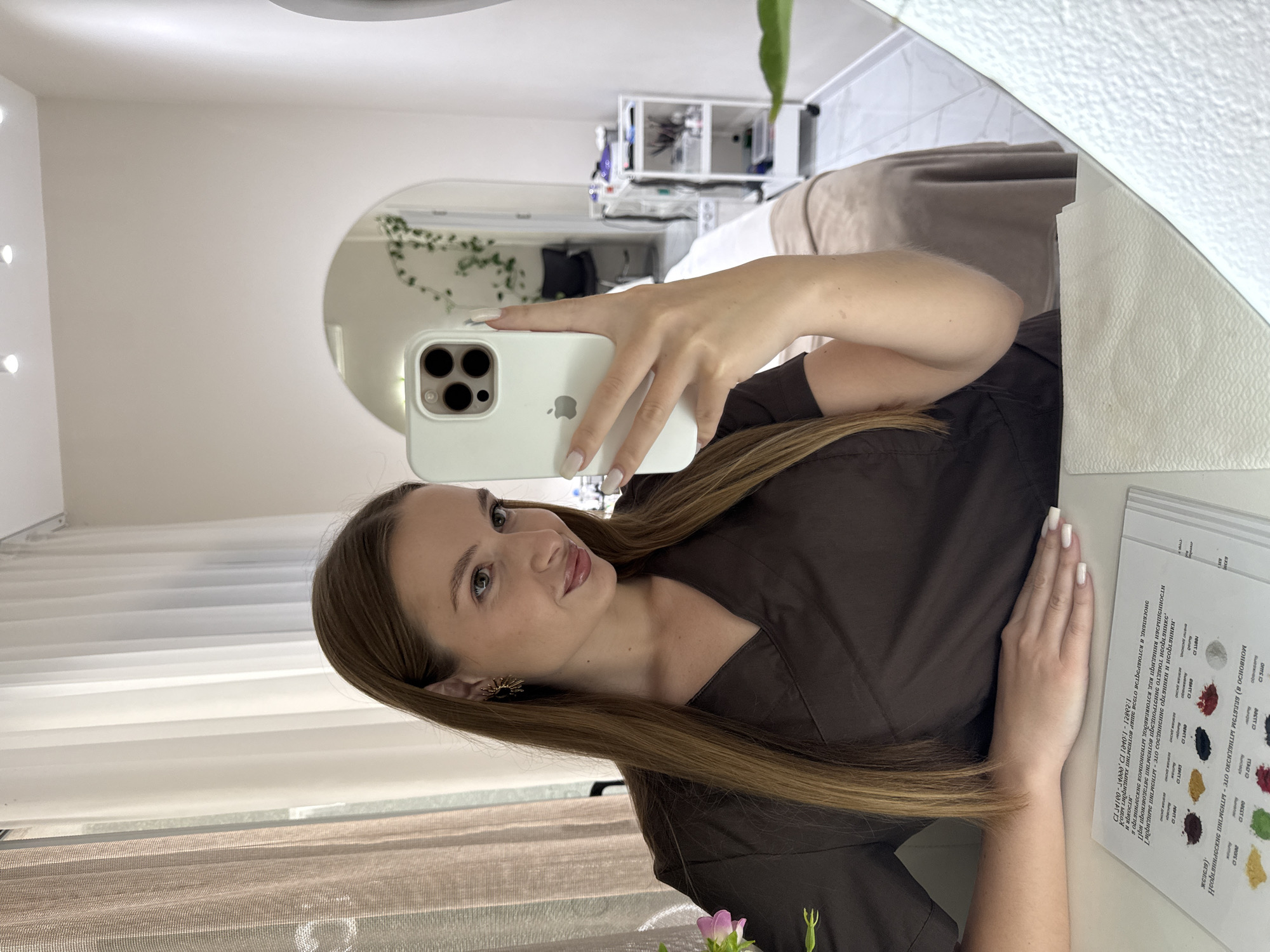 The image is a mirror selfie taken by a young woman in what appears to be a professional setting, likely a beauty salon or spa in Budva, Montenegro. The woman has long, straight brown hair and is wearing a dark brown, V-neck uniform top. She holds a white iPhone with a light-colored case, featuring a triple camera system and an Apple logo, capturing her reflection. Her fingernails are neatly painted white. She has a slight, pleasant smile, looking directly into the camera's lens, which is visible in her reflection.
The background, seen through the mirror, reveals a bright and clean room. White walls and what seems to be another decorative round mirror are visible in the distance. Shelving units stocked with various items, possibly salon supplies or products, are also reflected. A green plant or vine adds a subtle decorative element.
To the right of the woman's hand, a white sheet of paper is visible, covered with text in a Cyrillic script and multiple small, colored swatches or dots. This document appears to be a service menu, price list, or a catalog of product shades. While most of the text is blurred or small, some words are partially discernible, such as "ОСНОВНОЙ" (meaning "basic" or "main"), and various other terms like "SORTIO," "MAITIO," "Aumax," "DOSTT ID," and "ITASDIO," suggesting different categories or options. The overall impression is one of a well-maintained and professional establishment.