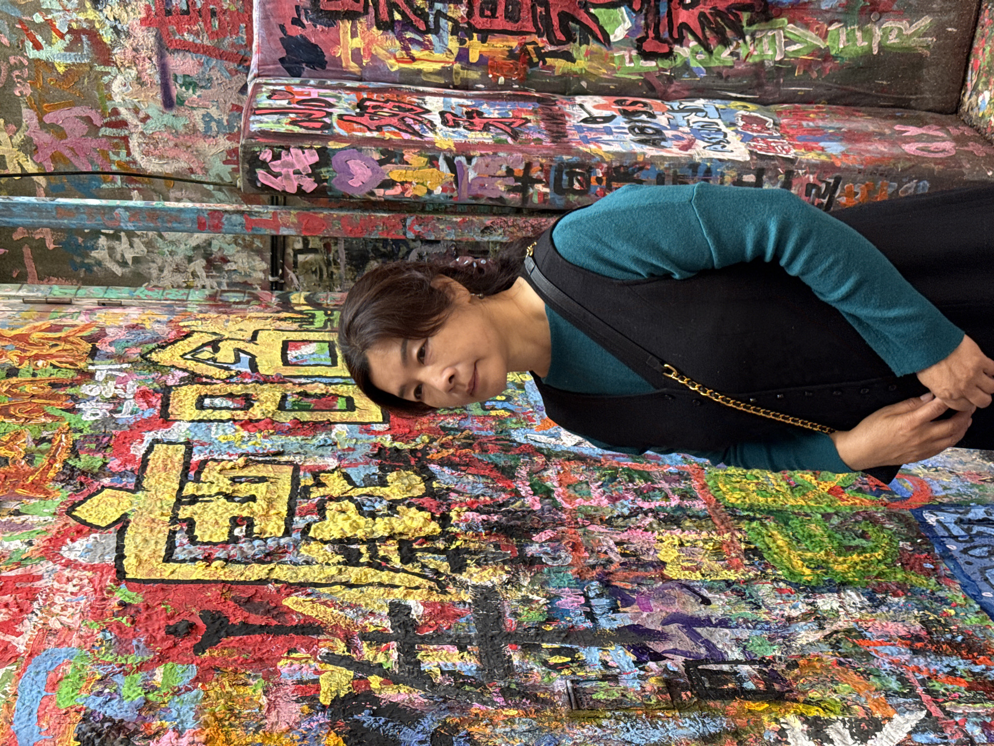 An adult female, appearing to be of East Asian descent, is depicted standing slightly right of center against a heavily graffitied background in Jiulongpo District, China. She gazes to her left with a neutral expression. Her dark hair is styled away from her face. She wears a long-sleeved teal garment underneath a black sleeveless outer layer, and a black shoulder bag with a gold chain strap is visible over her right shoulder. Her hands are clasped together at waist level. The surrounding environment consists of walls and vertical structures completely covered in thick, impasto street art. The graffiti displays a chaotic amalgamation of vibrant colors, including prominent reds, yellows, blues, greens, pinks, black, and white. Large yellow Chinese characters with black outlines are discernible on the left wall, interwoven with numerous smaller characters, abstract shapes, and the English word "ABYSS" visible on a column to the right. The paint application results in a highly textured, uneven surface.