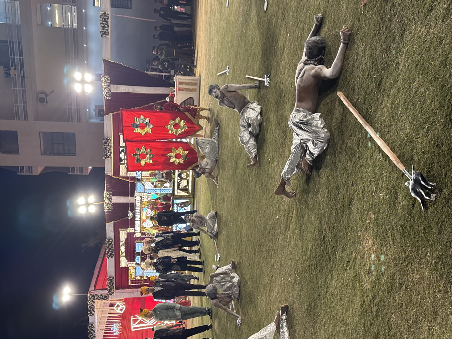 The image depicts a cultural celebration at night, possibly a wedding. People stand in formal attire near a decorated stage with red and gold banners. In the foreground, several performers are covered in white paint, wearing silver pants and lying on the grass. Some hold axes and a trident-like instrument. The scene blends traditional elements with theatrical performance.