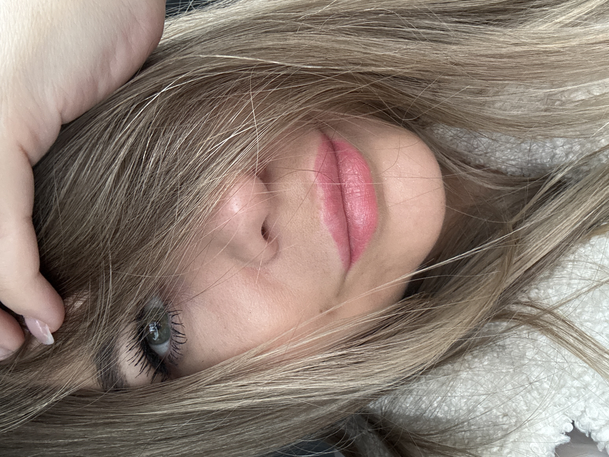 A close-up portrait depicts a female individual in Chaikovsky, Russia. Her long, light brown hair cascades over her forehead and the right side of her face, obscuring her right eye entirely. A light-skinned hand, exhibiting manicured fingernails with clear polish, is positioned at the top of her head, appearing to push back or adjust her hair. Her left eye is partially visible, revealing a green iris with prominently mascaraed upper lashes. The bridge and tip of her nose are discernible. Her lips are coated with bright pink lipstick and appear slightly parted. Her complexion is fair with visible skin texture. A white, textured garment, resembling faux fur or fleece, is visible on the lower left and bottom of the frame. The background is dark and out of focus, indicating an indeterminate interior or vehicle environment.