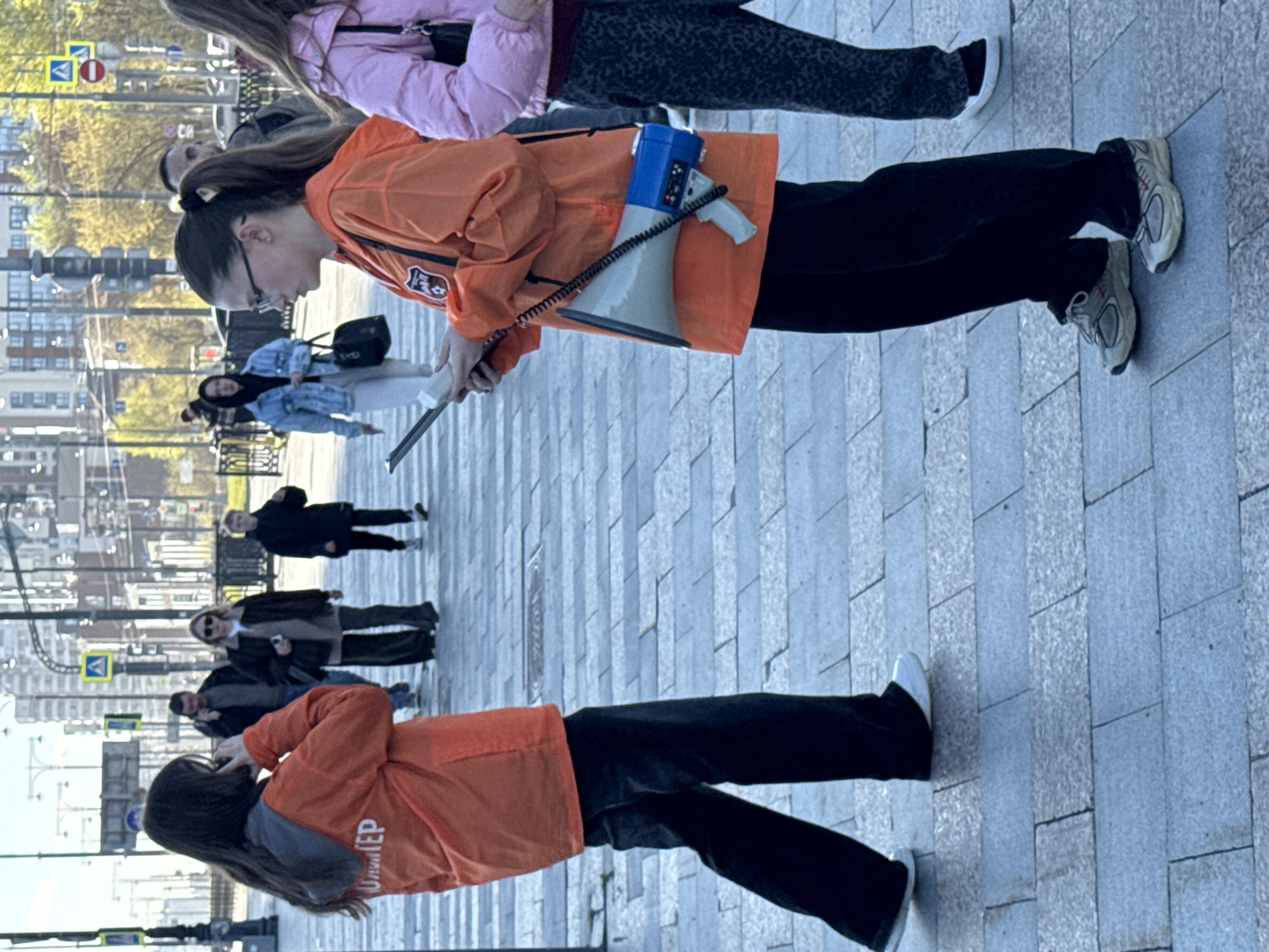 The image shows a group of people walking on a gray-paved street in Yekaterinburg, Russia. Two individuals are wearing bright orange jackets with text written on the back. One of them is holding a blue and gray megaphone. Other people are walking nearby, some in casual clothing such as jackets and jeans. Buildings and streetlights are visible in the background.