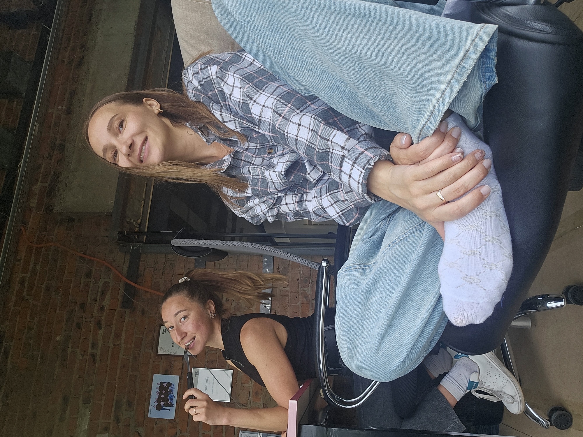 The image shows two women, likely in Chaikovsky, Russia given the Cyrillic script on posters in the background. The foreground features a woman with long brown hair, wearing a grey and white plaid shirt and light blue jeans, sitting on a black leather office chair. She is smiling at the camera and holding her foot. She has white socks. Behind her and to the left is another woman, also with brown hair pulled back in a ponytail, wearing a black tank top and sitting in another chair. She is holding scissors to her mouth and also smiling at the camera. The background includes a brick wall with several posters attached to it. The setting appears to be an office or workspace.