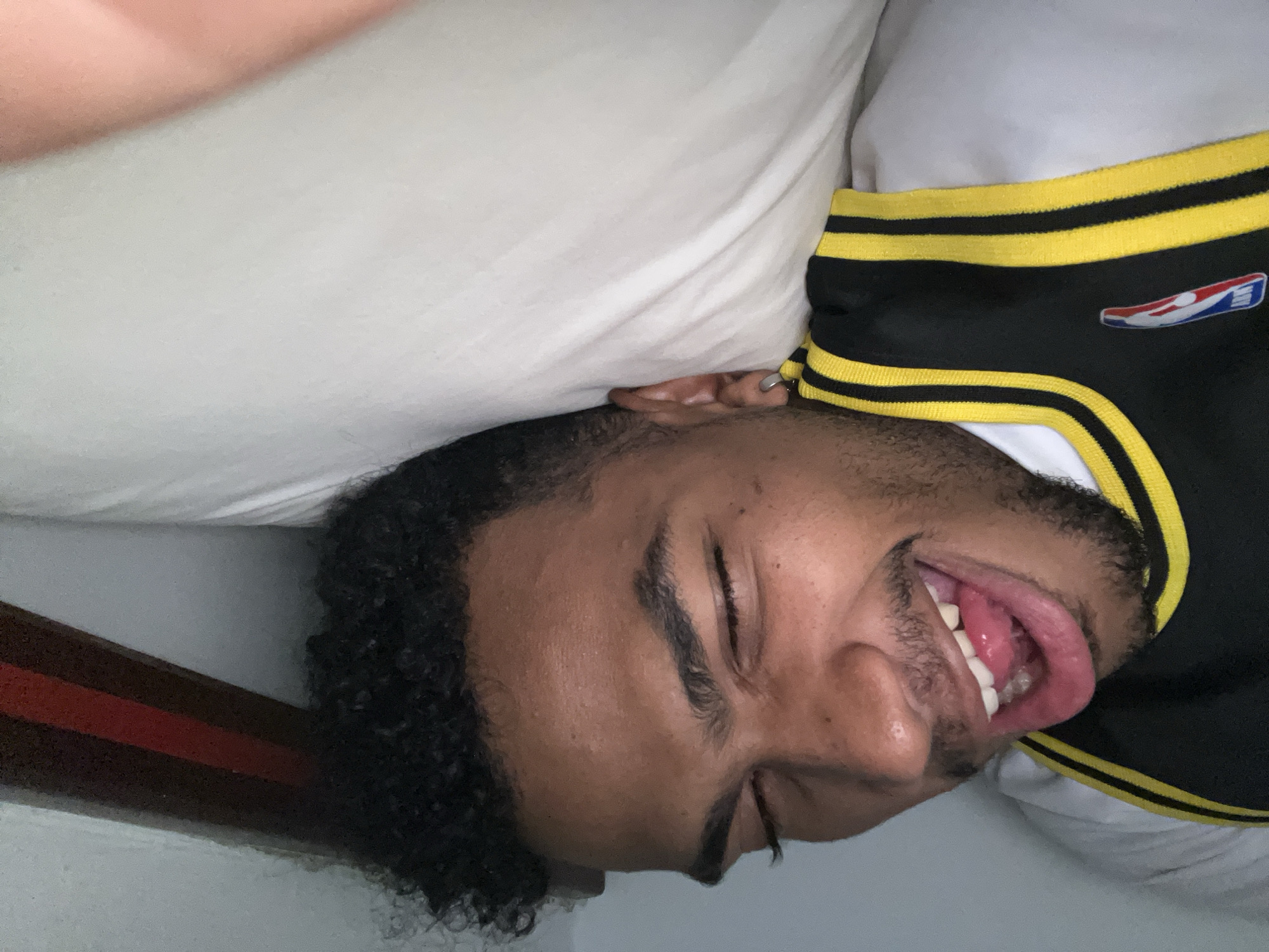 A person is lying on their side on a white pillow, wearing a black and yellow NBA jersey, with their tongue sticking out. The image is taken indoors. The location in the city of Porlamar, Venezuela cannot be confirmed from the given image.